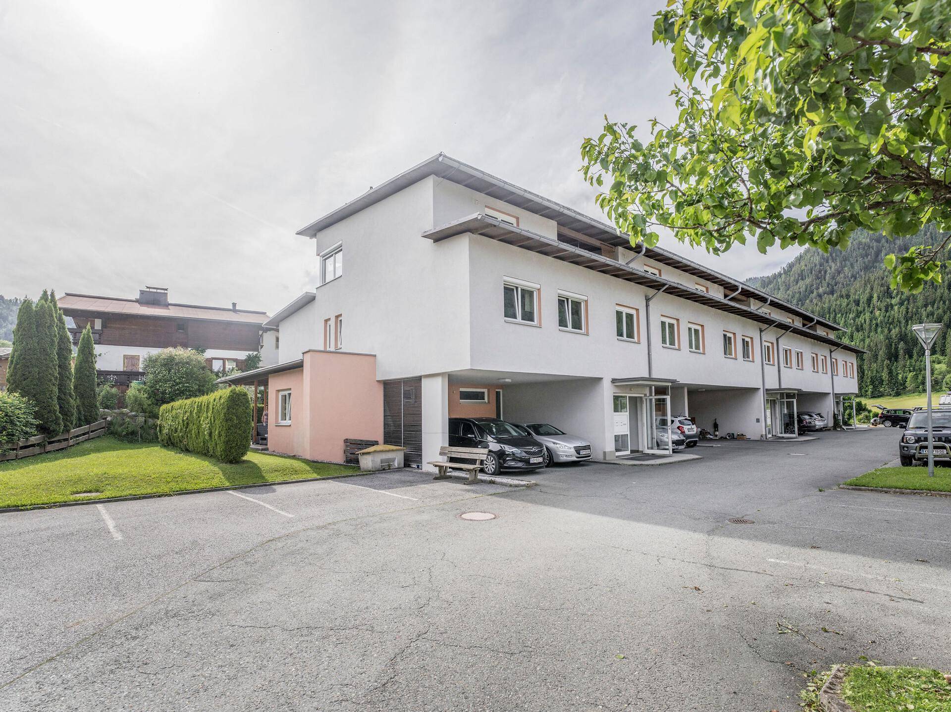 Modernes Mehrfamilienhaus mit Parkplätzen und grüner Umgebung vor einer Bergkulisse.