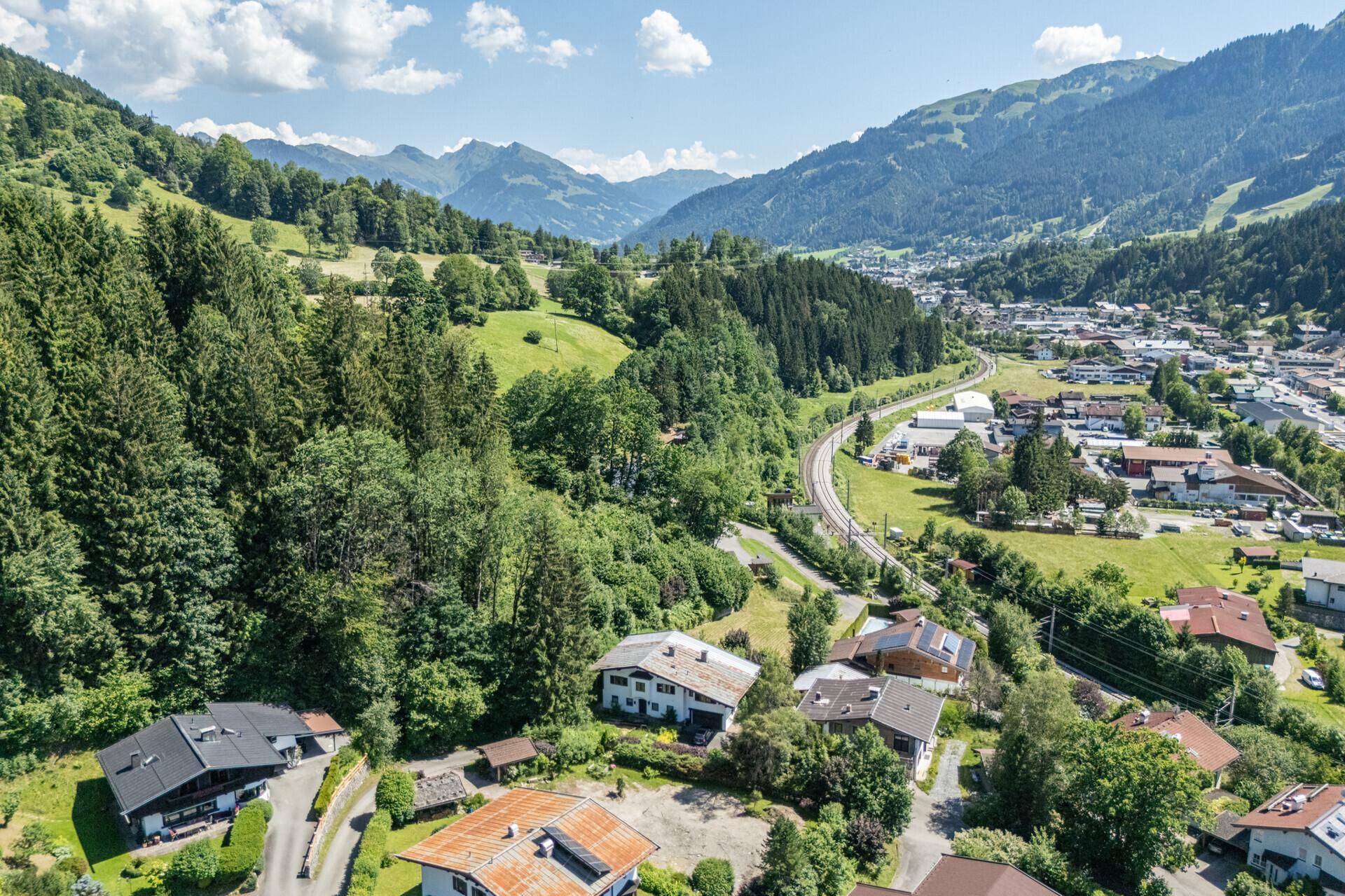 Panoramablick auf ein Bergtal mit einer Wohnsiedlung, umgeben von Wäldern und einer Bahnlinie.