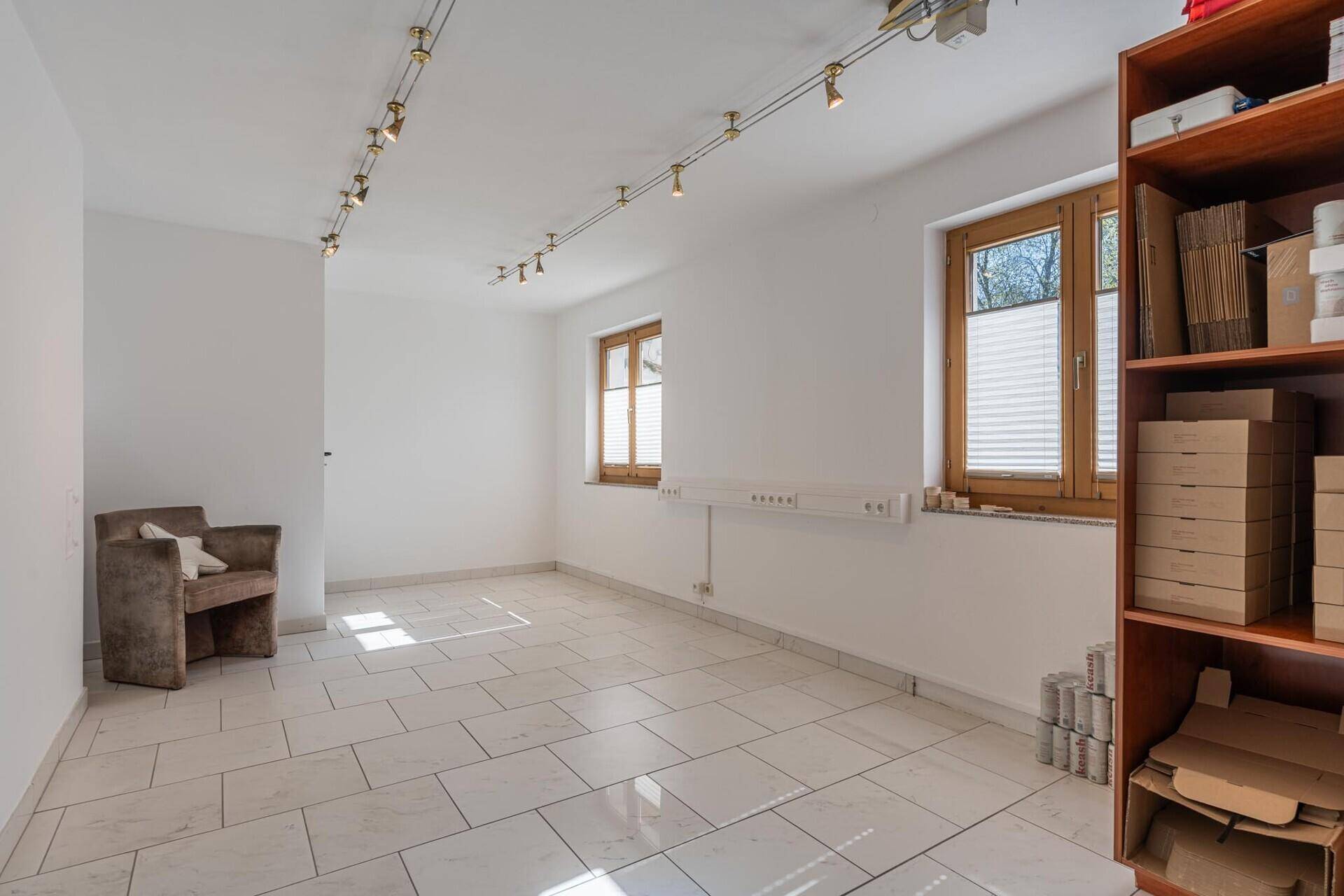 Heller Mehrzweckraum mit Fliesenboden, Regalen, zwei Fenstern und viel Tageslicht.