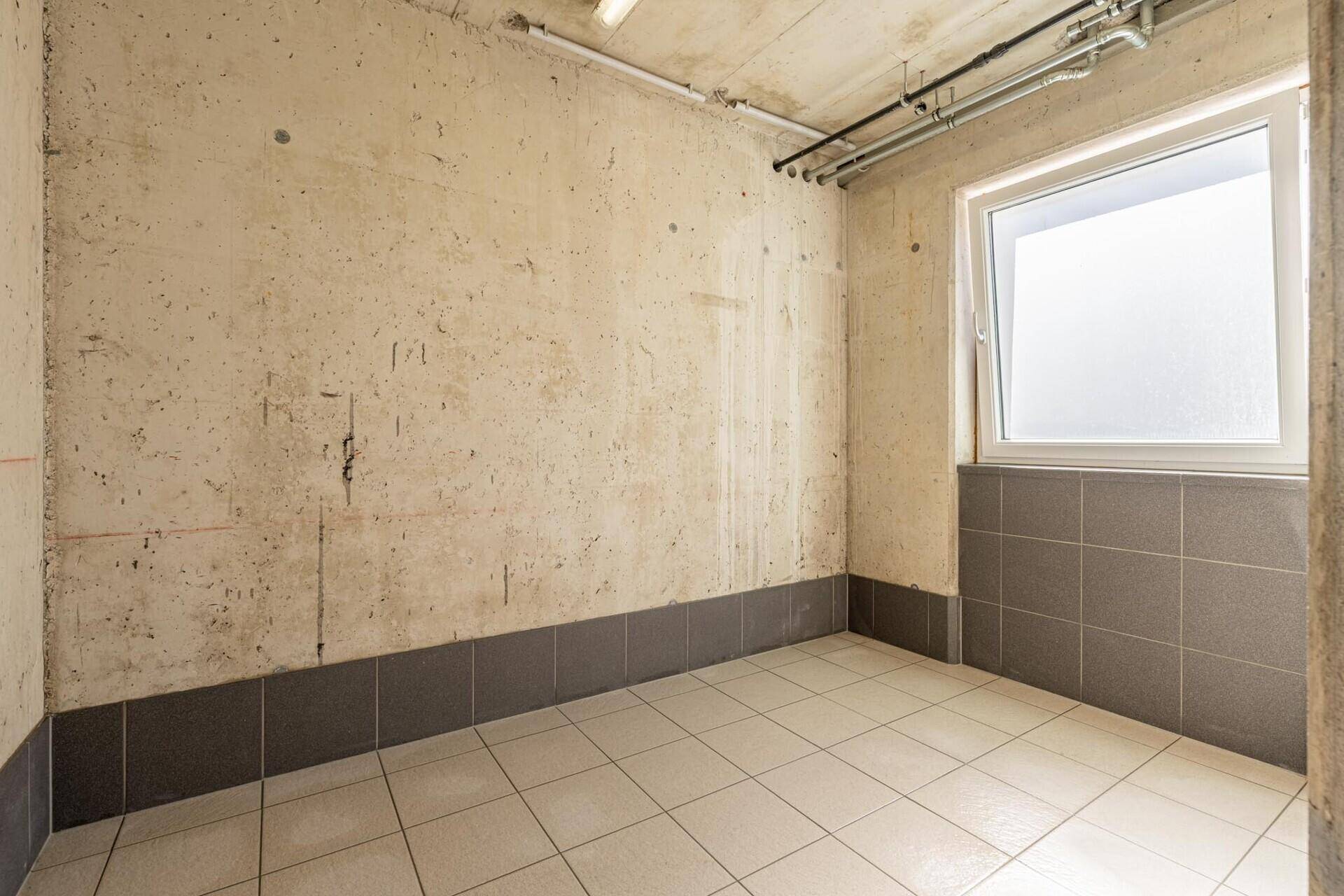 Kleiner Raum mit Betonwänden, Fliesenboden und einem Fenster für natürliches Licht.