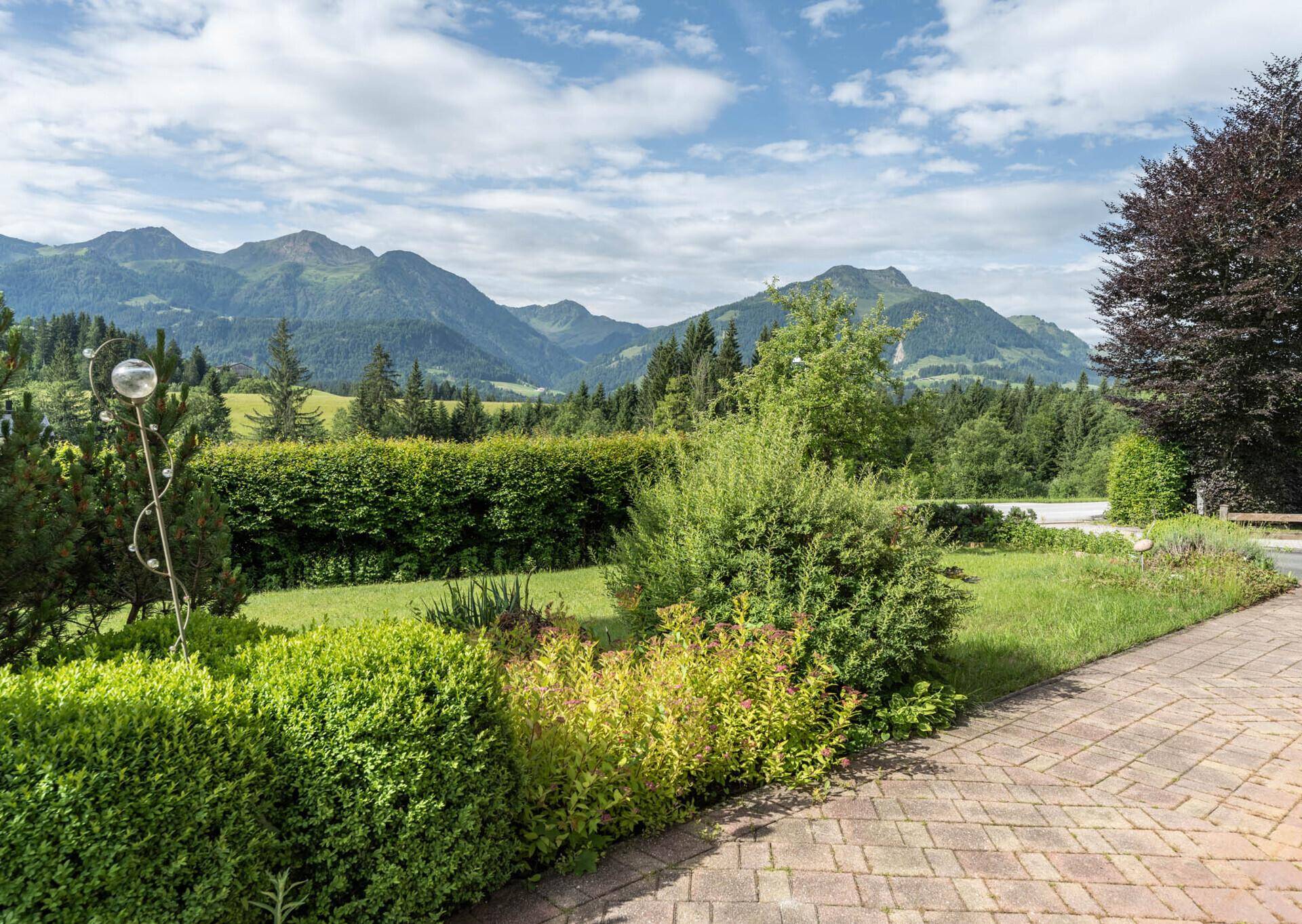 Gepflegter Gartenbereich mit Sträuchern, gepflastertem Weg und beeindruckendem Bergblick.
