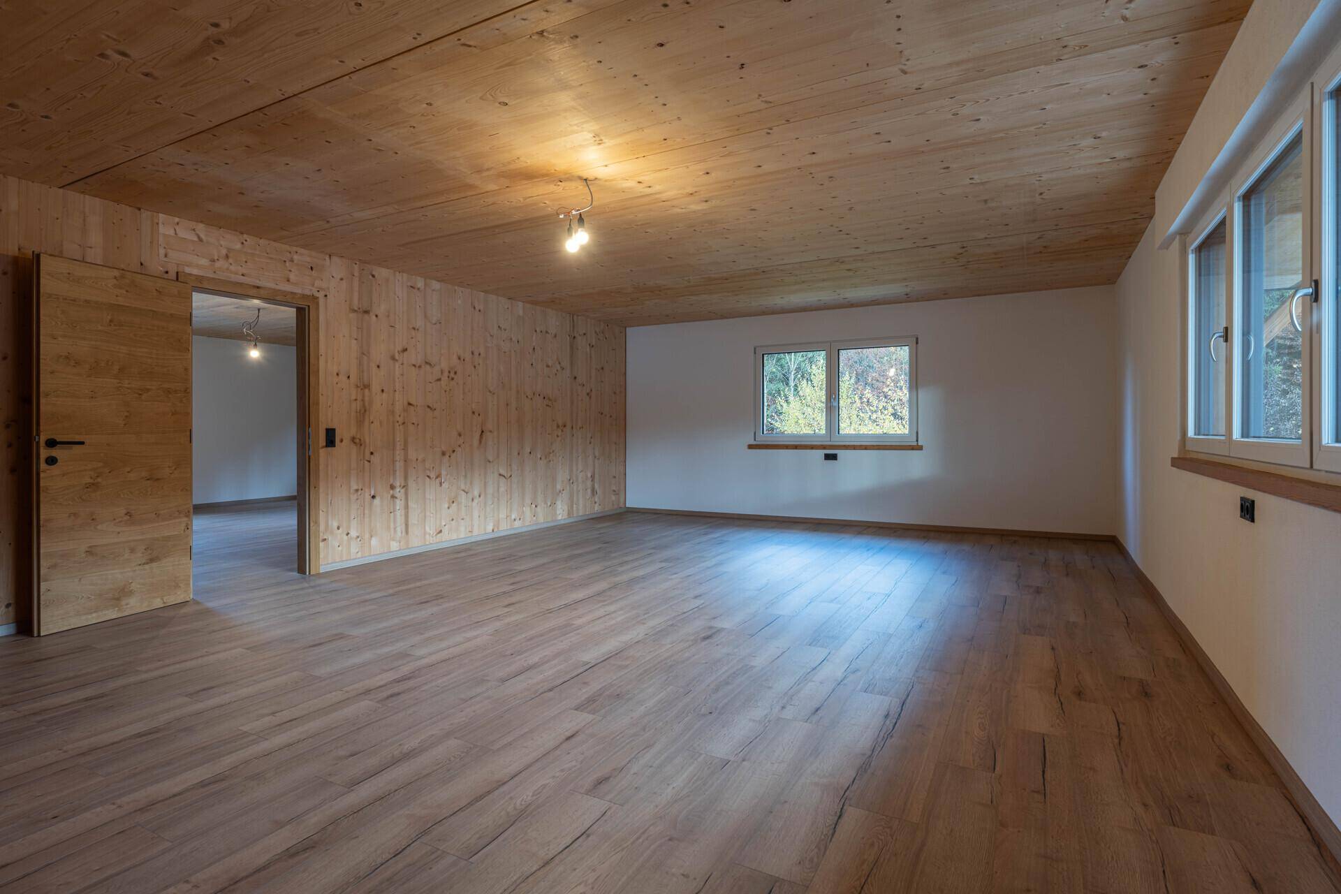 Heller, leerer Raum mit Holzwänden, Holzdecke und großen Fenstern, die viel Tageslicht hereinlassen.