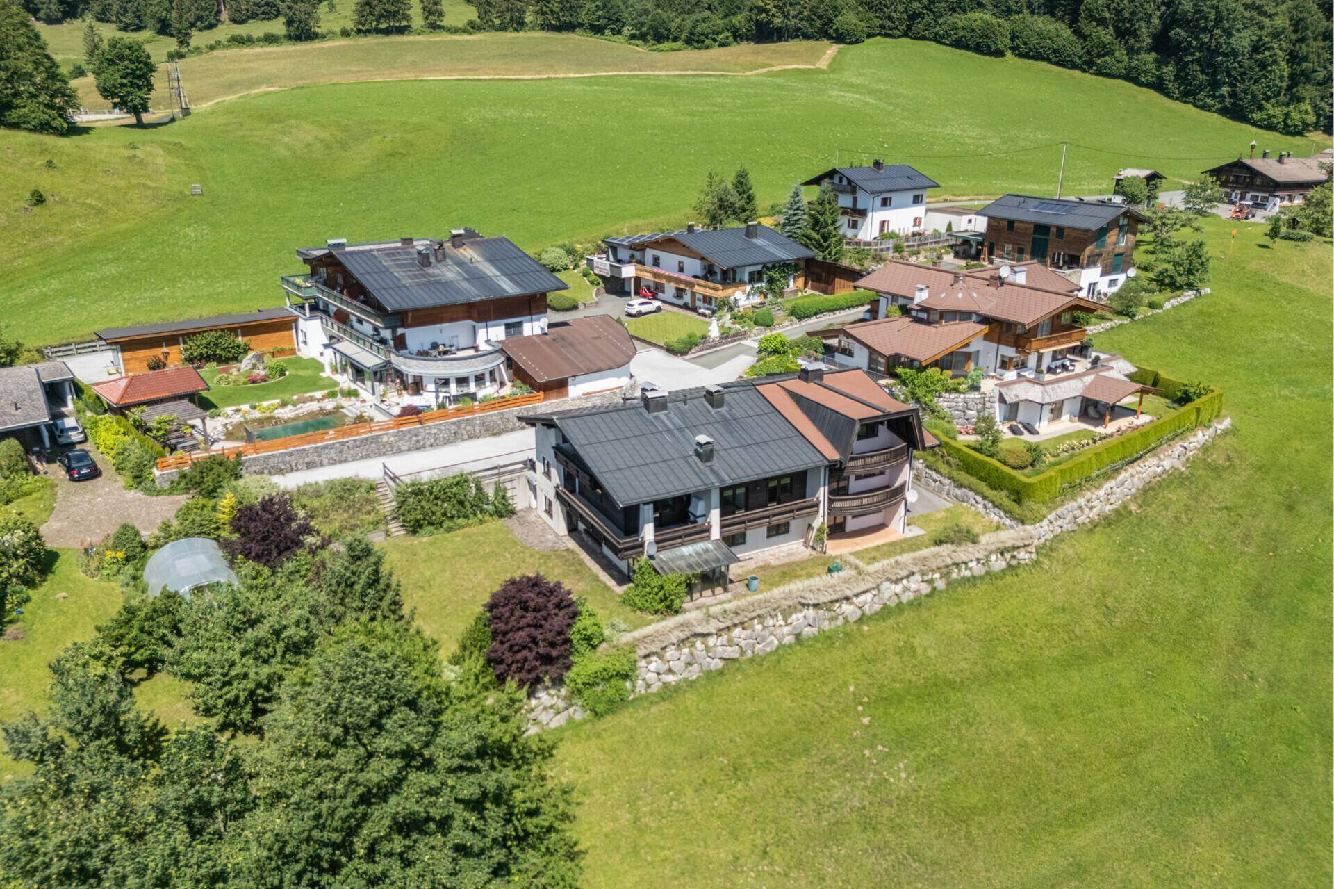 Luftaufnahme einer Wohnsiedlung mit mehreren Häusern, umgeben von grünen Wiesen und Bäumen, unter blauem Himmel.