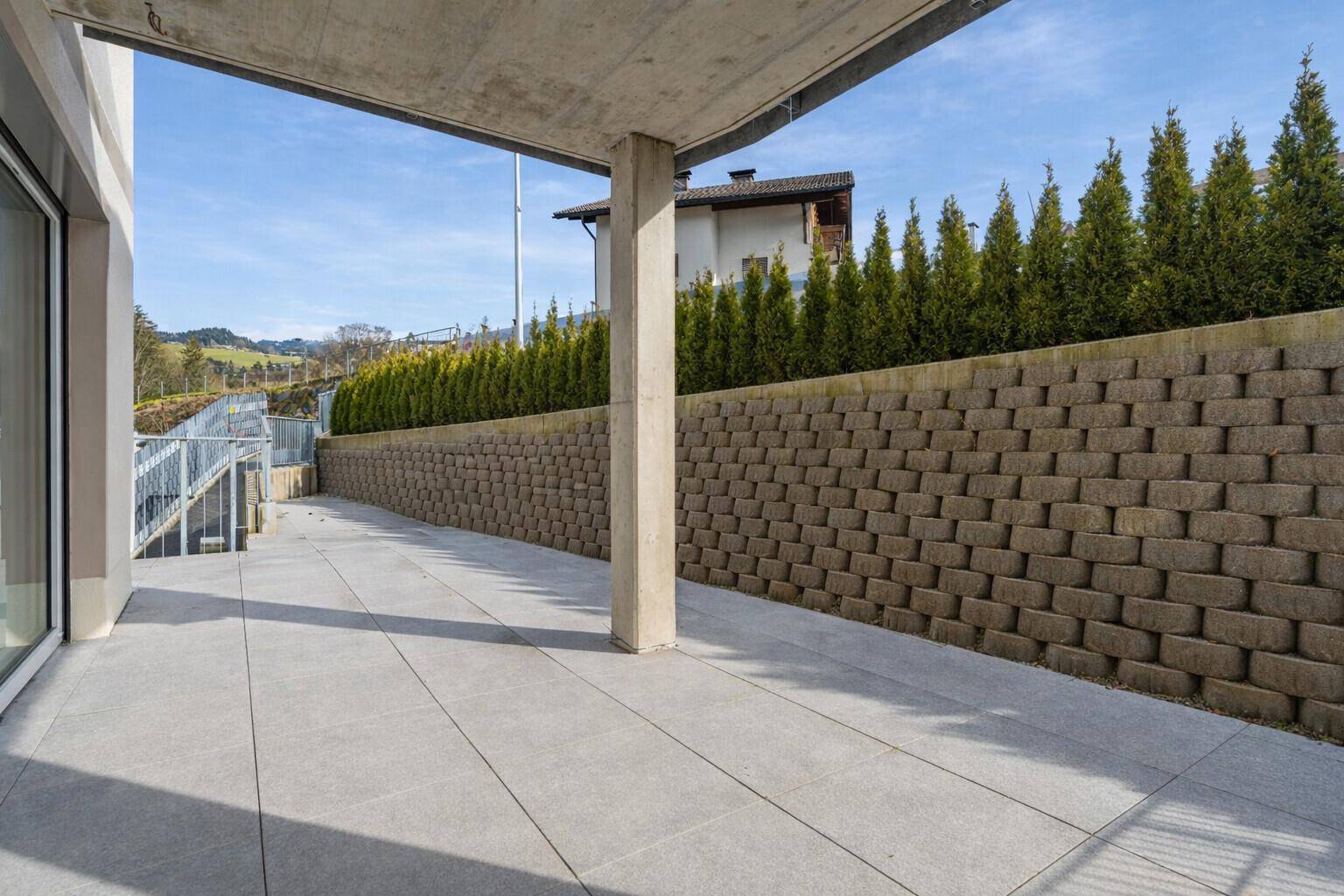 Geräumige, geflieste Terrasse mit einer hohen Stützmauer und einer grünen Hecke im Hintergrund.