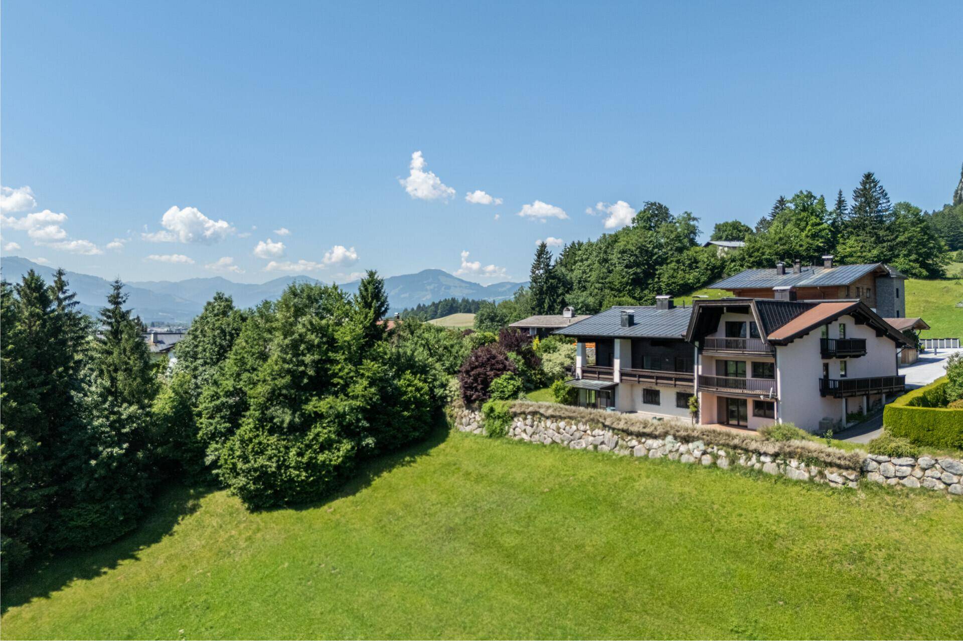 Erhöhter Blick auf Wohnhäuser in einer grünen Umgebung mit bewaldeten Hügeln und Bergen am Horizont.