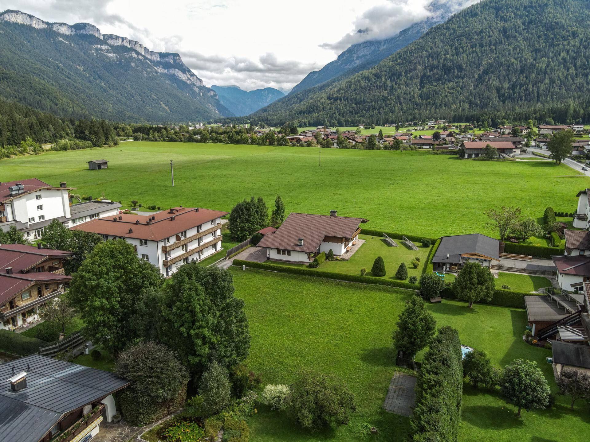 Panoramablick auf ein weitläufiges grünes Grundstück, umgeben von einem Dorf und imposanten Bergketten.