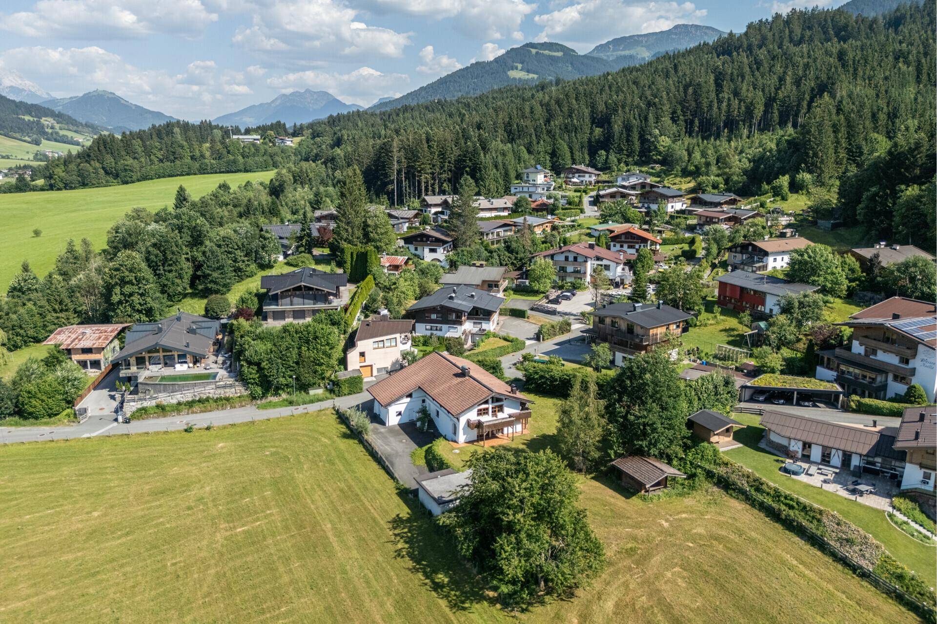 Panoramablick auf eine ländliche Wohnsiedlung mit grünen Wiesen und bewaldeten Hügeln im Hintergrund.
