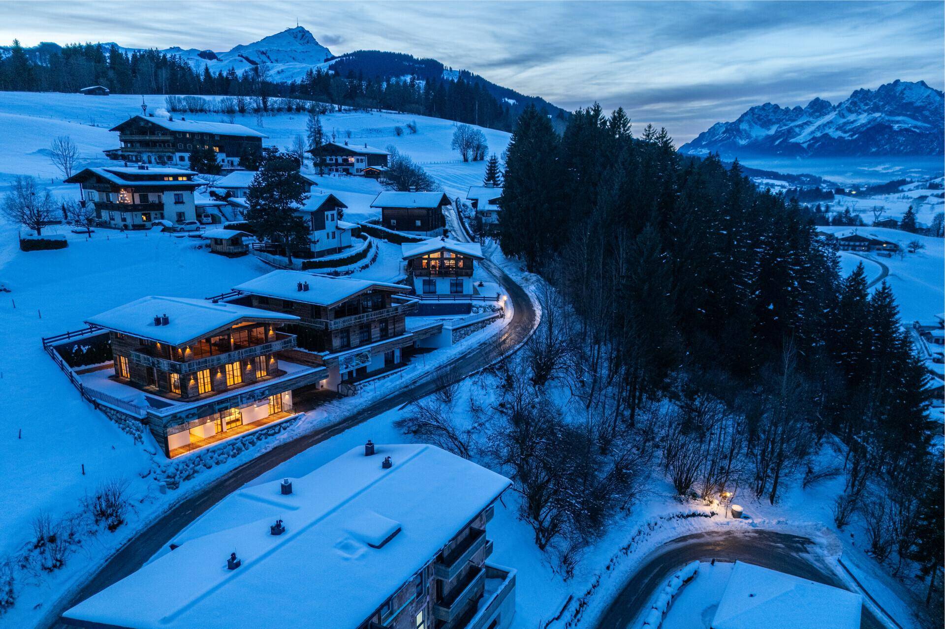 Panoramablick auf eine verschneite Berglandschaft mit mehreren luxuriösen Chalets bei Dämmerung.