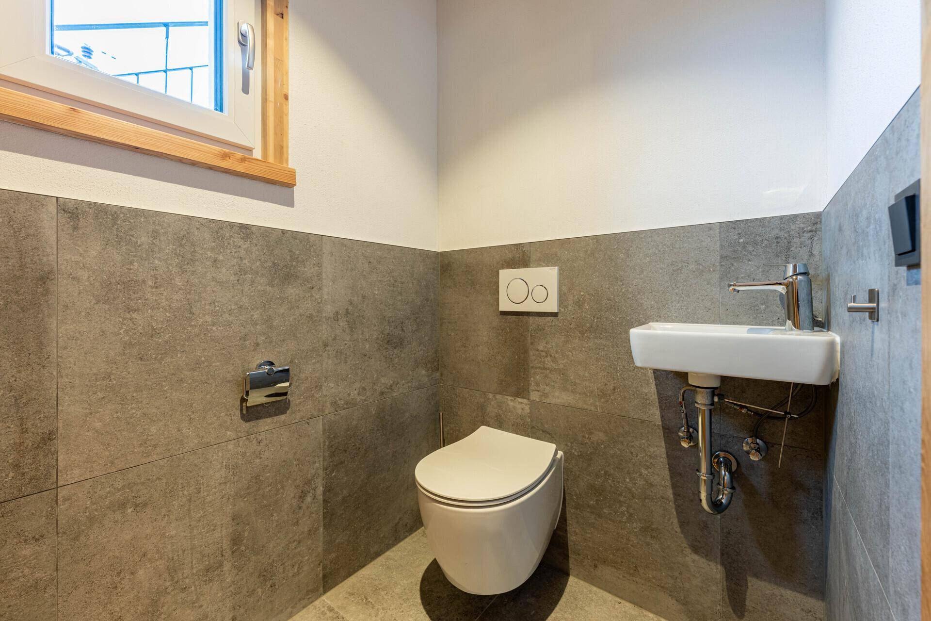 Modernes Gäste-WC mit grauen Fliesen, wandhängender Toilette, kleinem Waschbecken und Fenster.