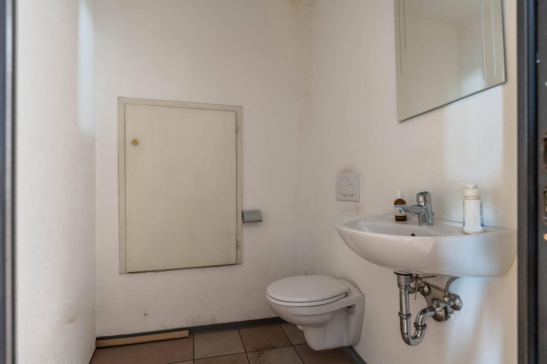 Einfache Gästetoilette mit wandhängendem WC, Waschbecken und einem kleinen Spiegel.