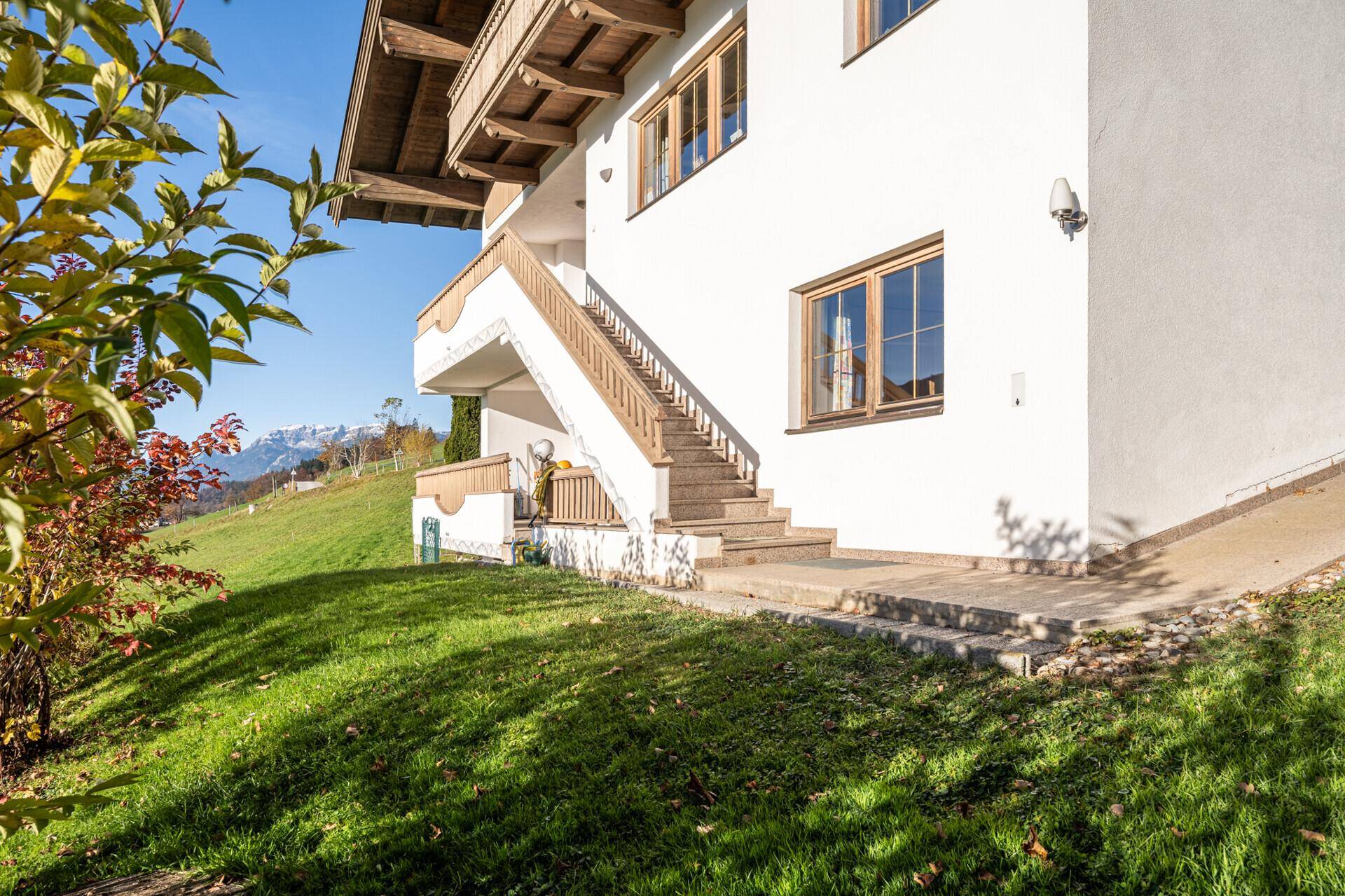 Weiße Hausfassade mit Holzfenstern und Außentreppe, umgeben von einer grünen Rasenfläche.
