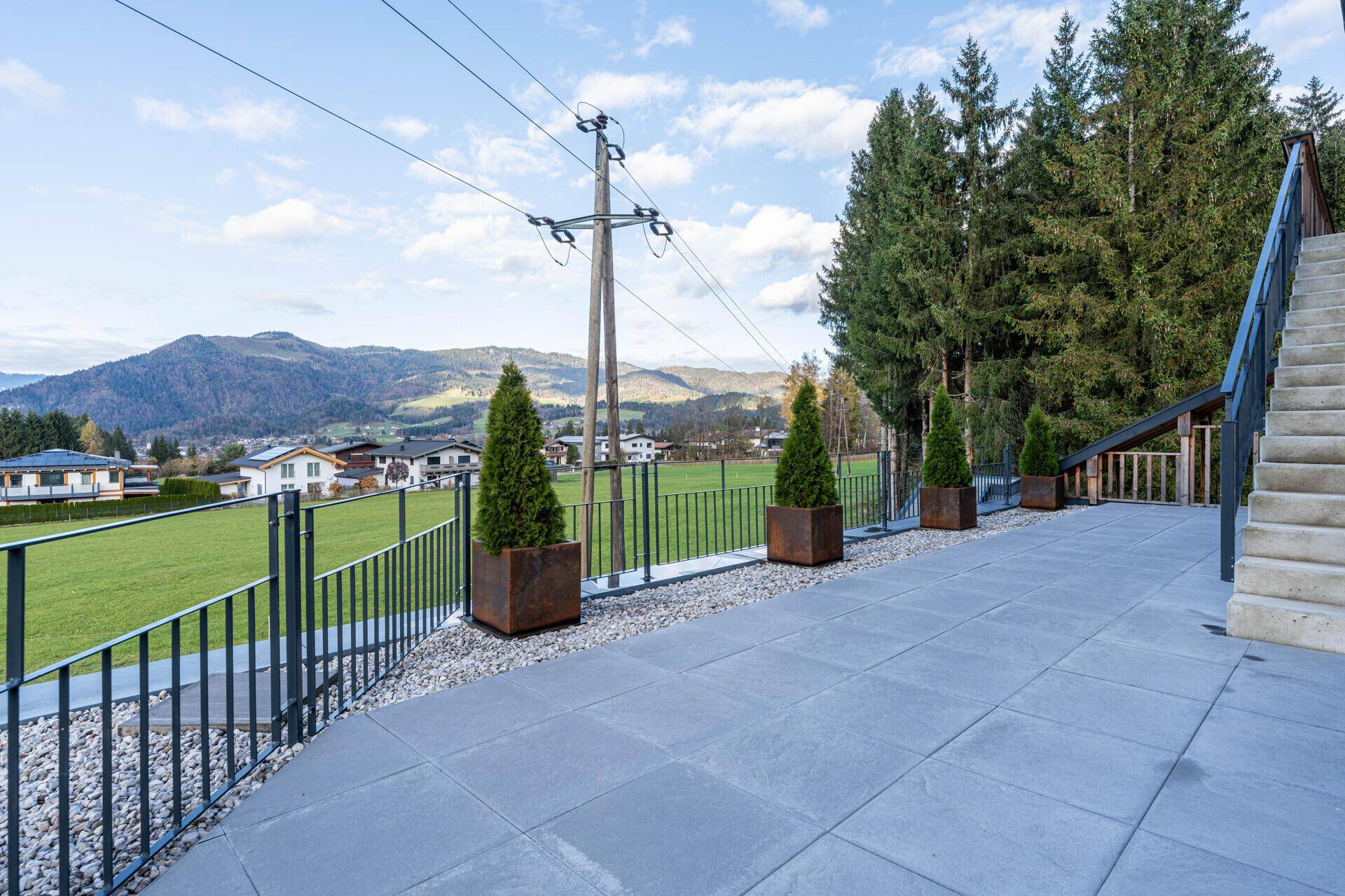 Gepflasterte Terrasse mit Blick auf grüne Hügel und Berge, umgeben von Bäumen und einem Zaun.