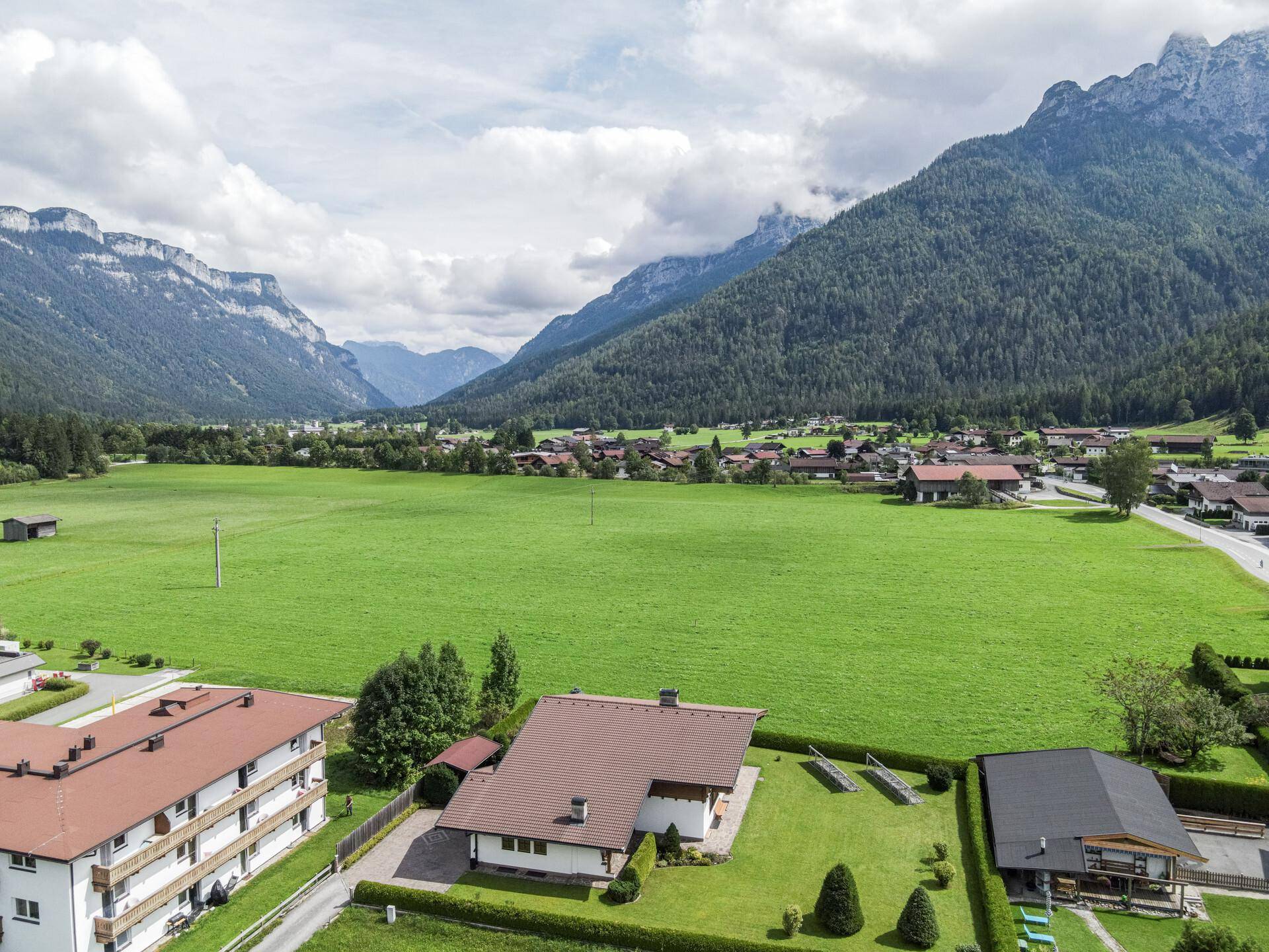 Weitläufige Ansicht eines grünen Grundstücks mit Dorfbebauung und majestätischen Bergen im Hintergrund.
