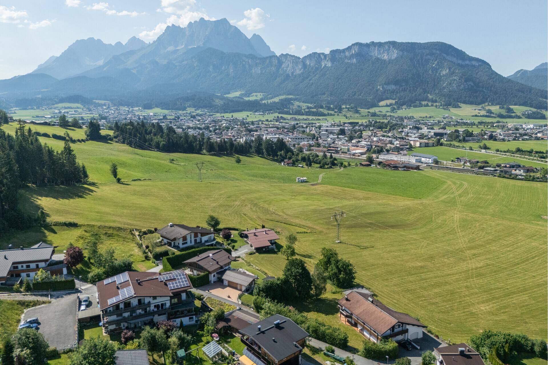 Weitläufige Luftaufnahme einer Siedlung mit vielen Häusern, grünen Feldern und einer beeindruckenden Bergkulisse.