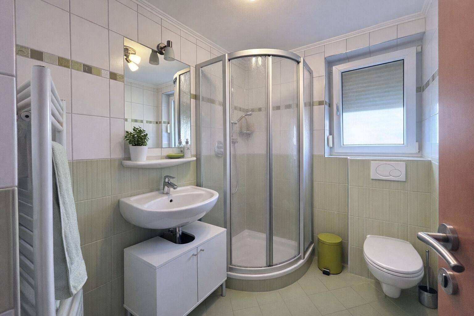 Helles Badezimmer mit Duschkabine, Fenster und grün-weißen Fliesen.