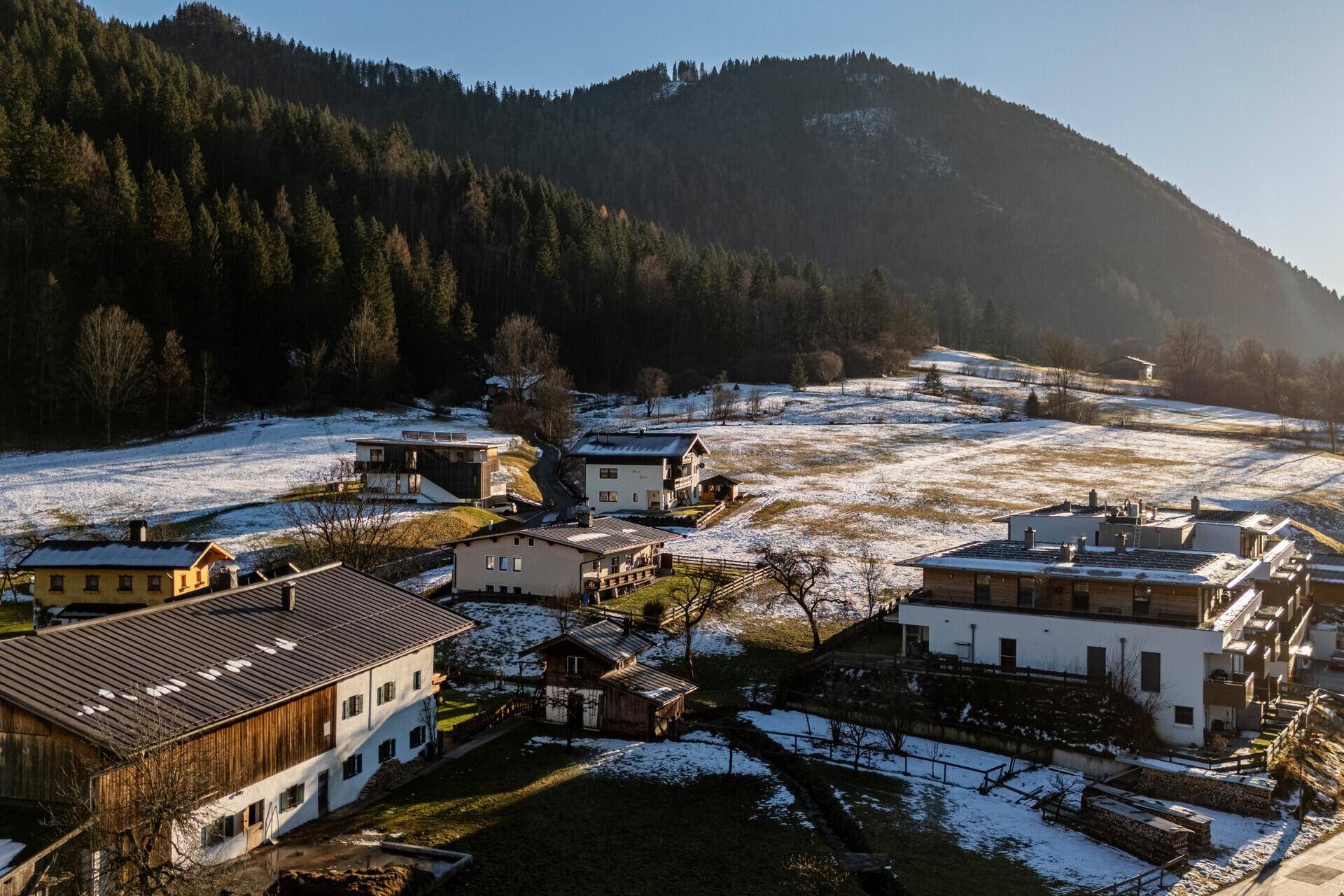 Luftaufnahme einer Siedlung mit Häusern auf einem schneebedeckten Hang, umgeben von Wald und Bergen.