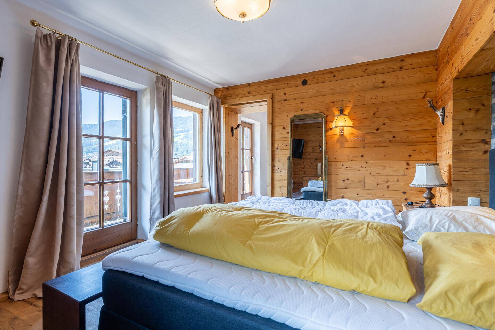 Helles Schlafzimmer mit Holzwand, Doppelbett und Zugang zu einem kleinen Balkon mit Bergblick.