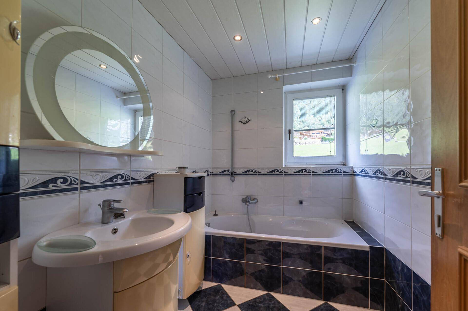 Badezimmer mit Badewanne, Fenster und hellen Fliesen in Weiß und Blau im älteren Stil.