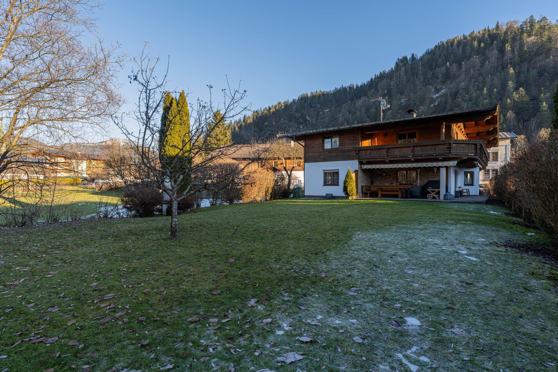 Großer Gartenbereich mit Rasenfläche und einem traditionellen Holzhaus im alpinen Stil, umgeben von Bergen.
