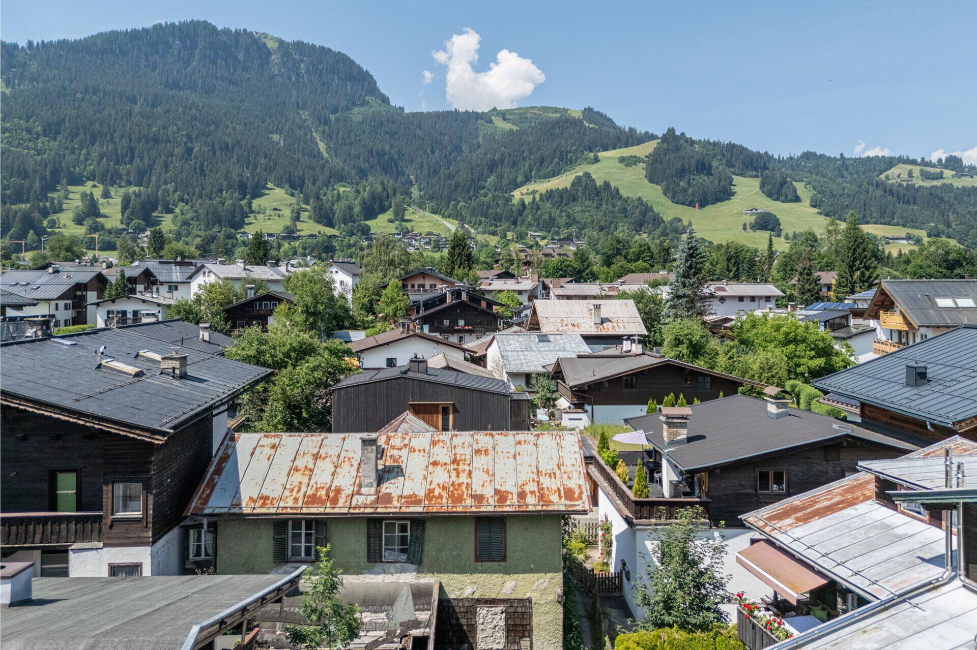 Blick auf ein traditionelles Haus mit rostigem Dach und einer beeindruckenden Bergkulisse unter blauem Himmel.