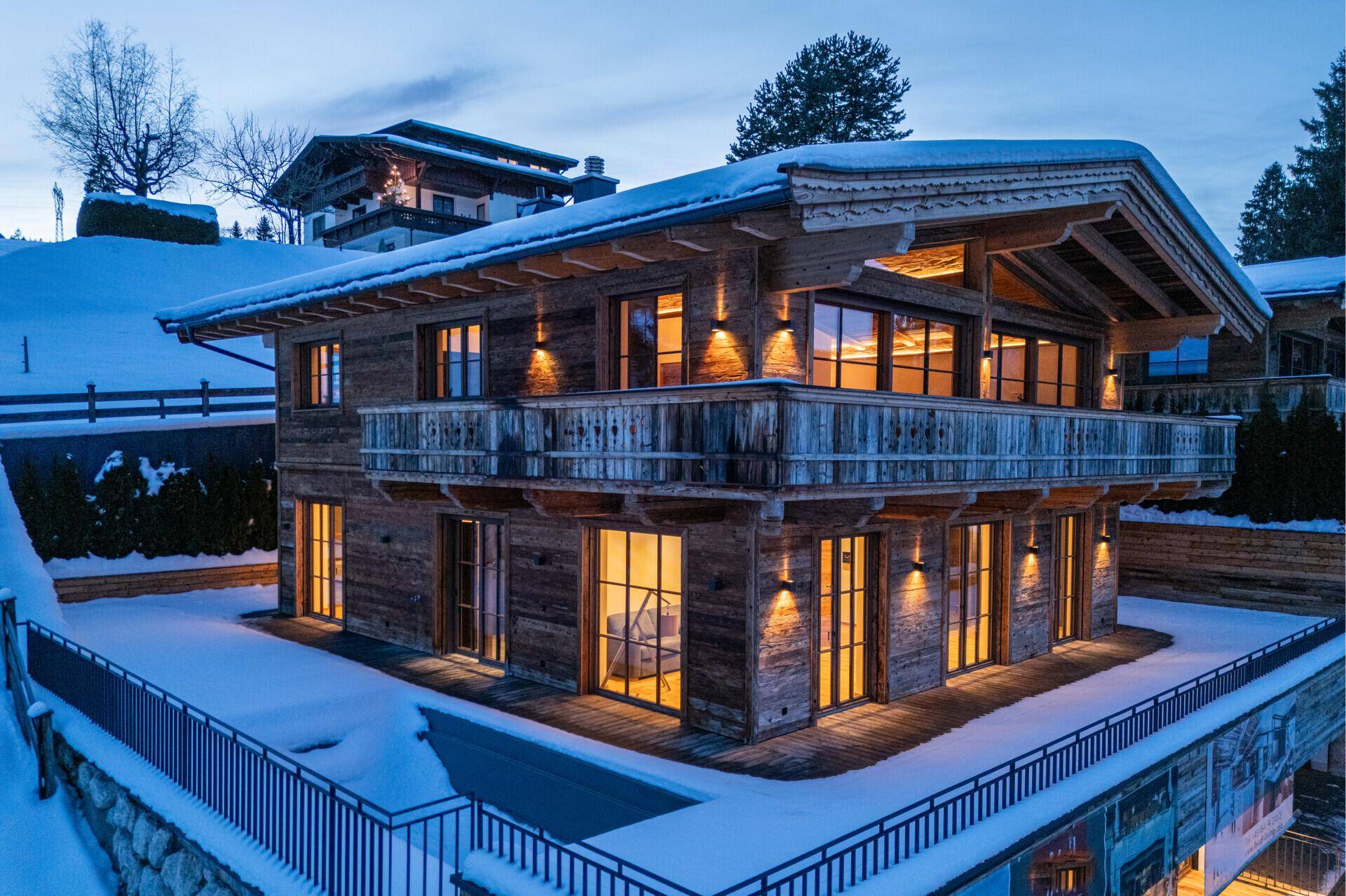 Elegantes Holzchalet in verschneiter Umgebung, mit beleuchteten Fenstern und Balkon bei Abenddämmerung.