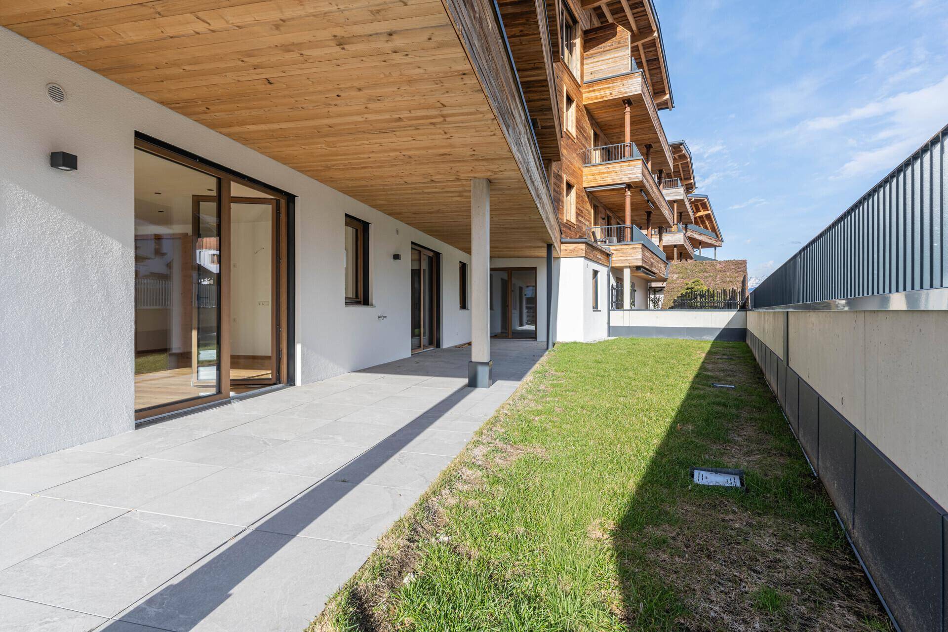 Lange, gepflasterte Terrasse entlang des Gebäudes mit Rasenfläche und Bergblick.