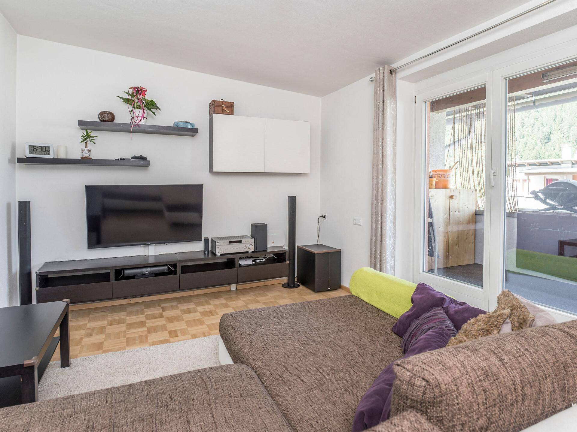 Wohnzimmer mit Sofa, Fernseher und Zugang zu einem Balkon, hell durch große Glastüren.