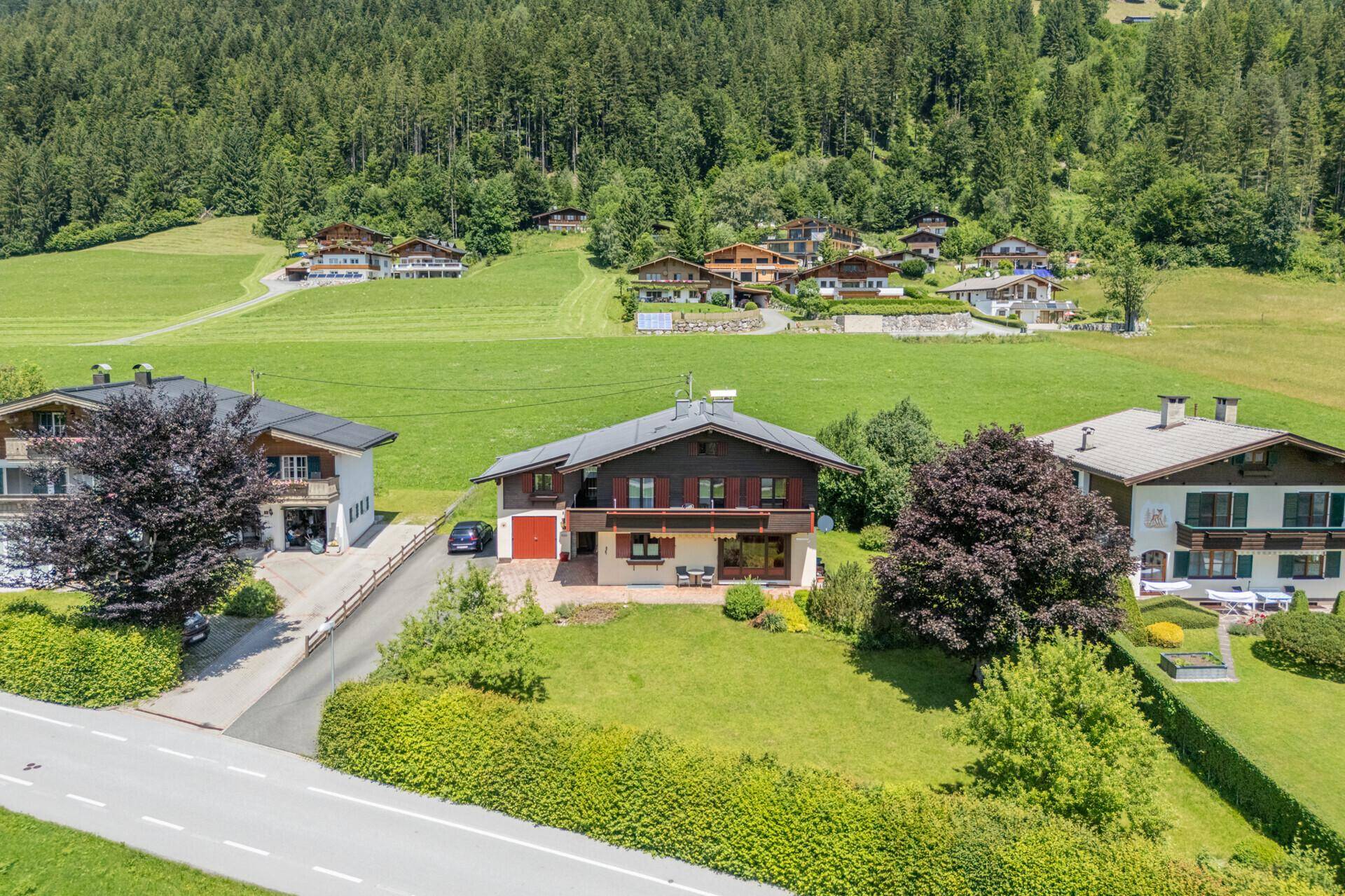 Haus mit roter Garage und Terrasse, eingebettet in eine grüne Landschaft mit bewaldeten Hügeln und Bergen.