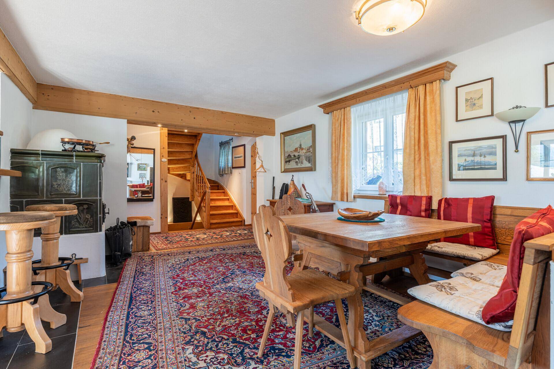 Traditioneller Essbereich mit Holztisch, Eckbank und Kachelofen, verbunden mit einer Holztreppe.