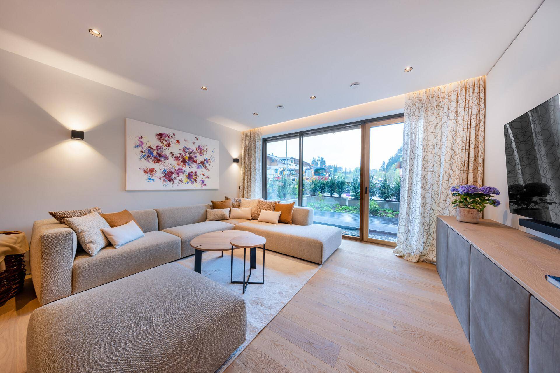 Helles Wohnzimmer mit bequemer Eckcouch, Holzboden und großen Fenstern mit Blick ins Grüne.