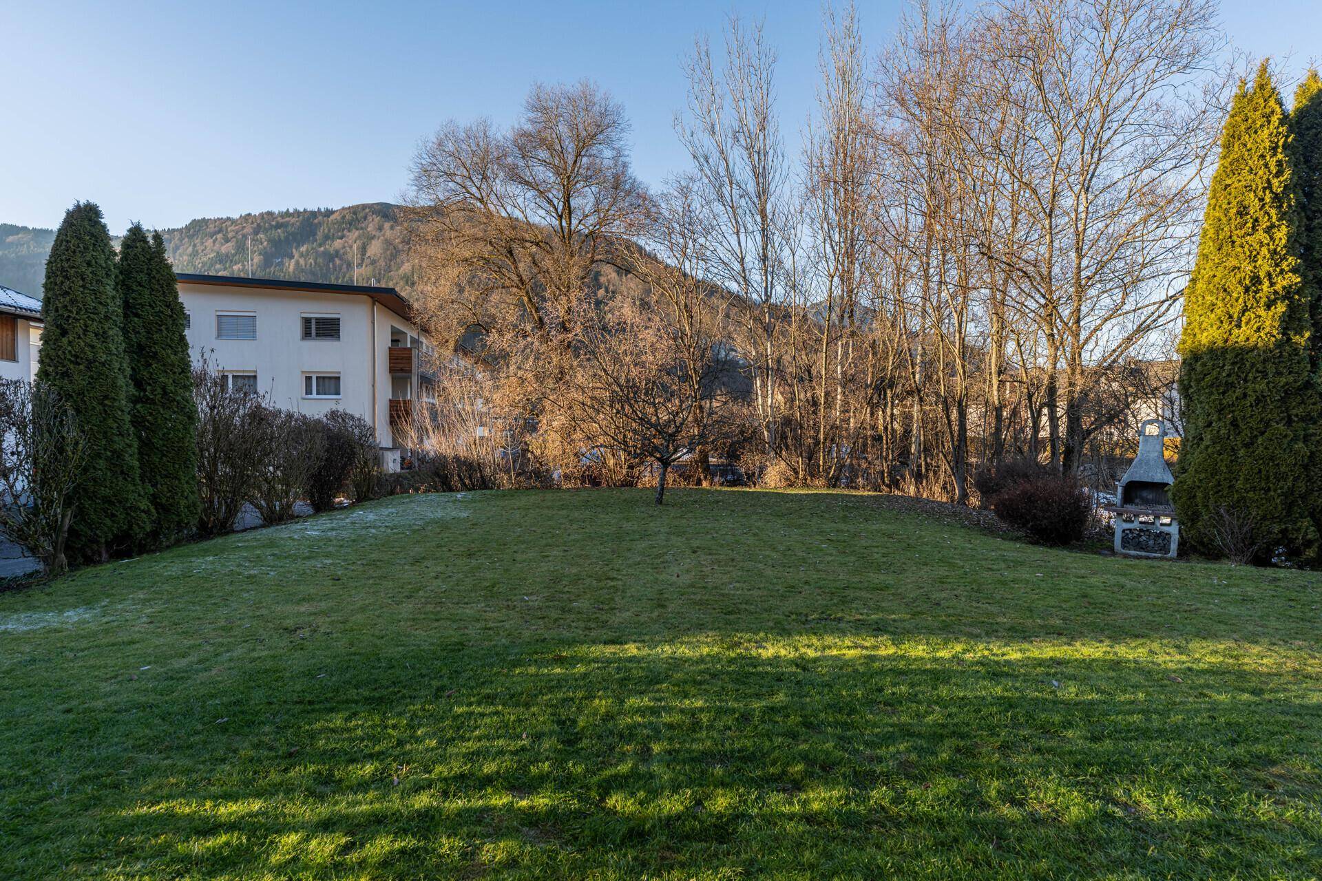 Gepflegter Garten mit Rasenfläche, Grill und Blick auf die umliegenden Berge und Gebäude.