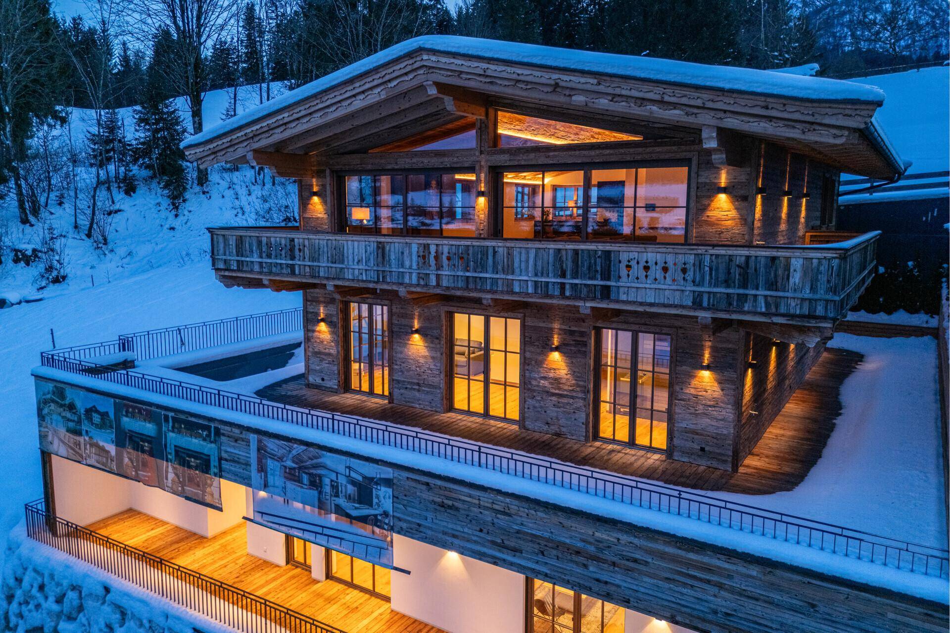 Luxuriöses Chalet in den Bergen bei Dämmerung, mit beleuchteten Balkonen und Terrassen im Schnee.