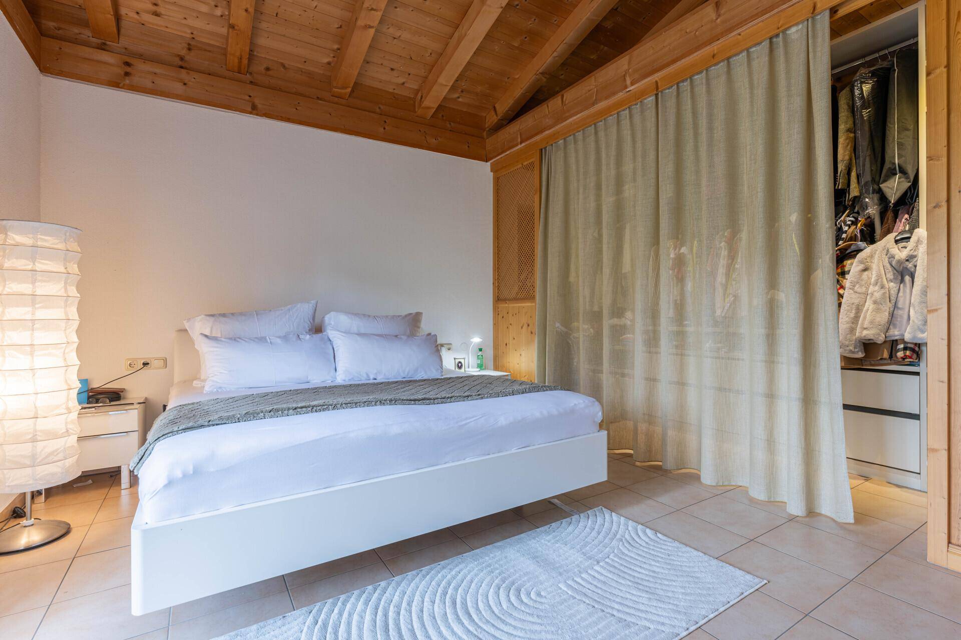 Schlafzimmer mit Fliesenboden, schwebendem Bett und begehbarem Kleiderschrank hinter Vorhang.