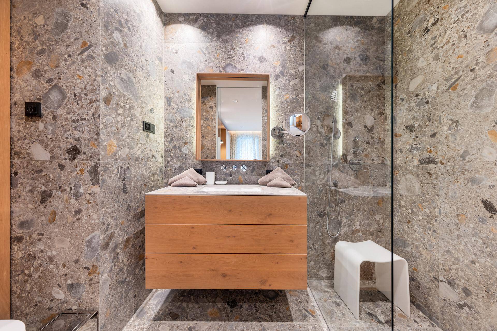 Modernes Badezimmer mit Steinfliesen, schwebendem Holzwaschtisch und beleuchtetem Spiegel.