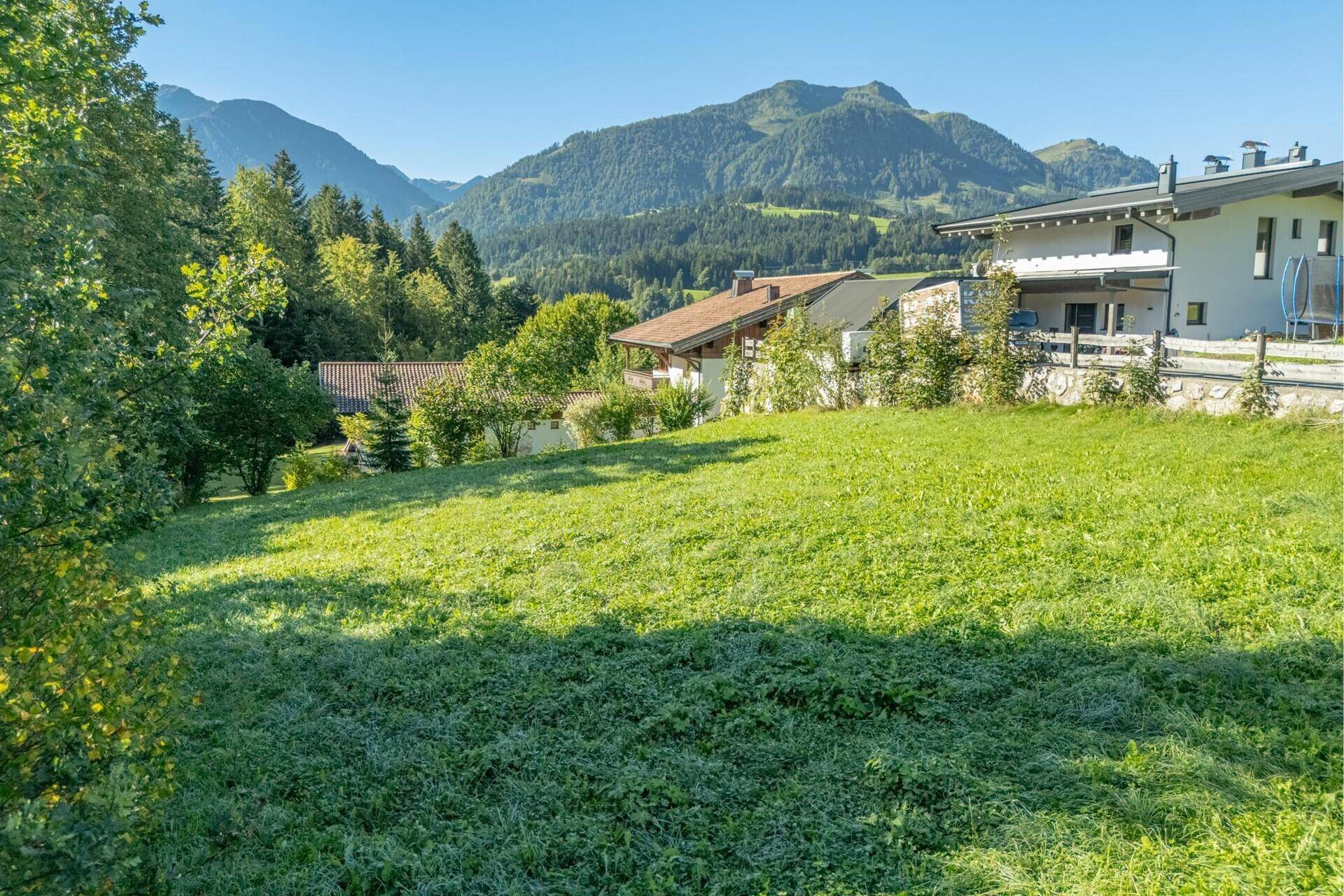 Sonnige Rasenfläche mit leichtem Gefälle und Häusern im Hintergrund, umgeben von Bergen.