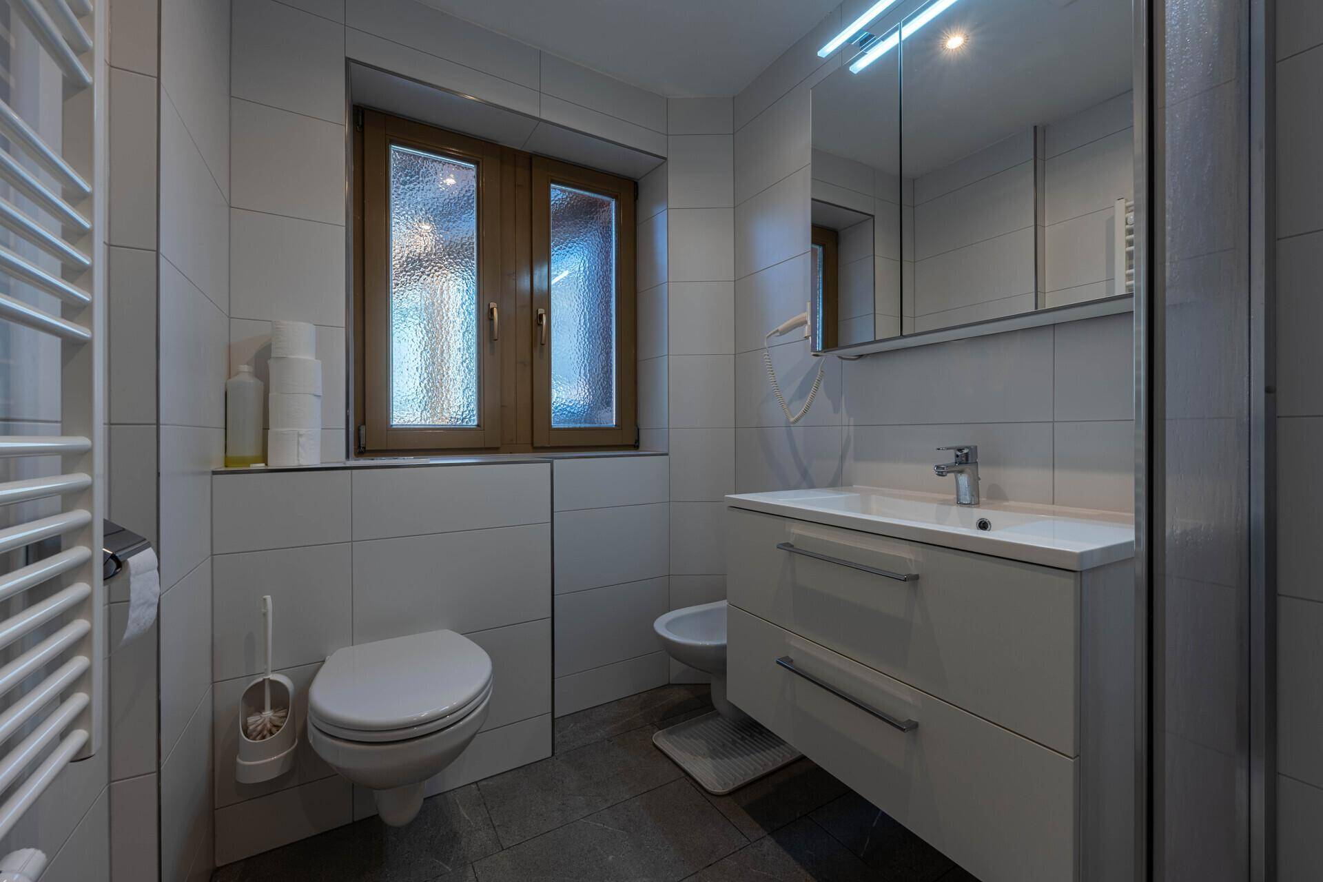 Modernes Badezimmer mit weißen Fliesen, Fenster, WC, Bidet und Waschtisch mit Spiegelschrank.