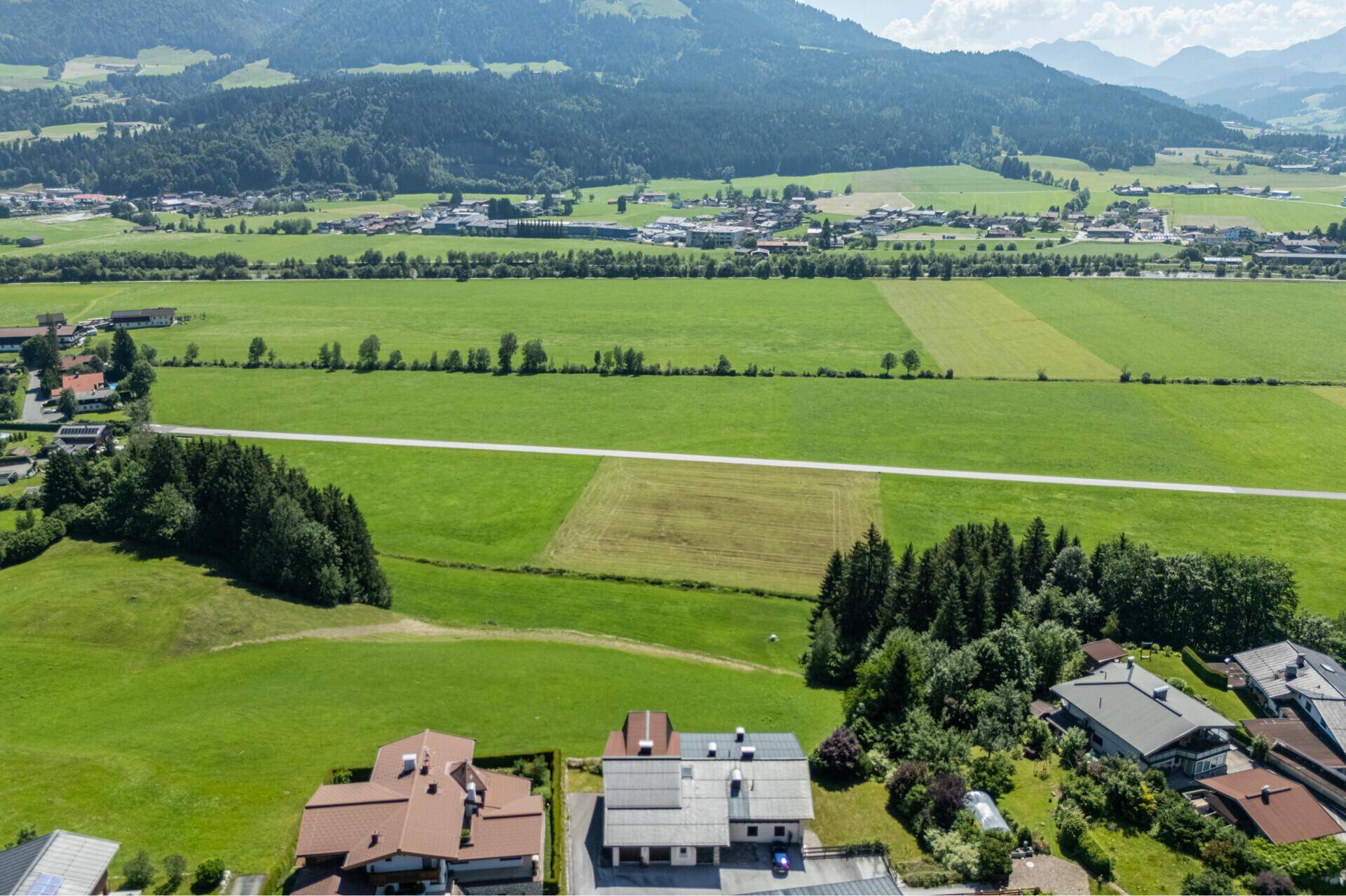 Weitläufige grüne Felder und Berge umgeben eine kleine Wohnsiedlung mit Häusern und Solaranlagen.