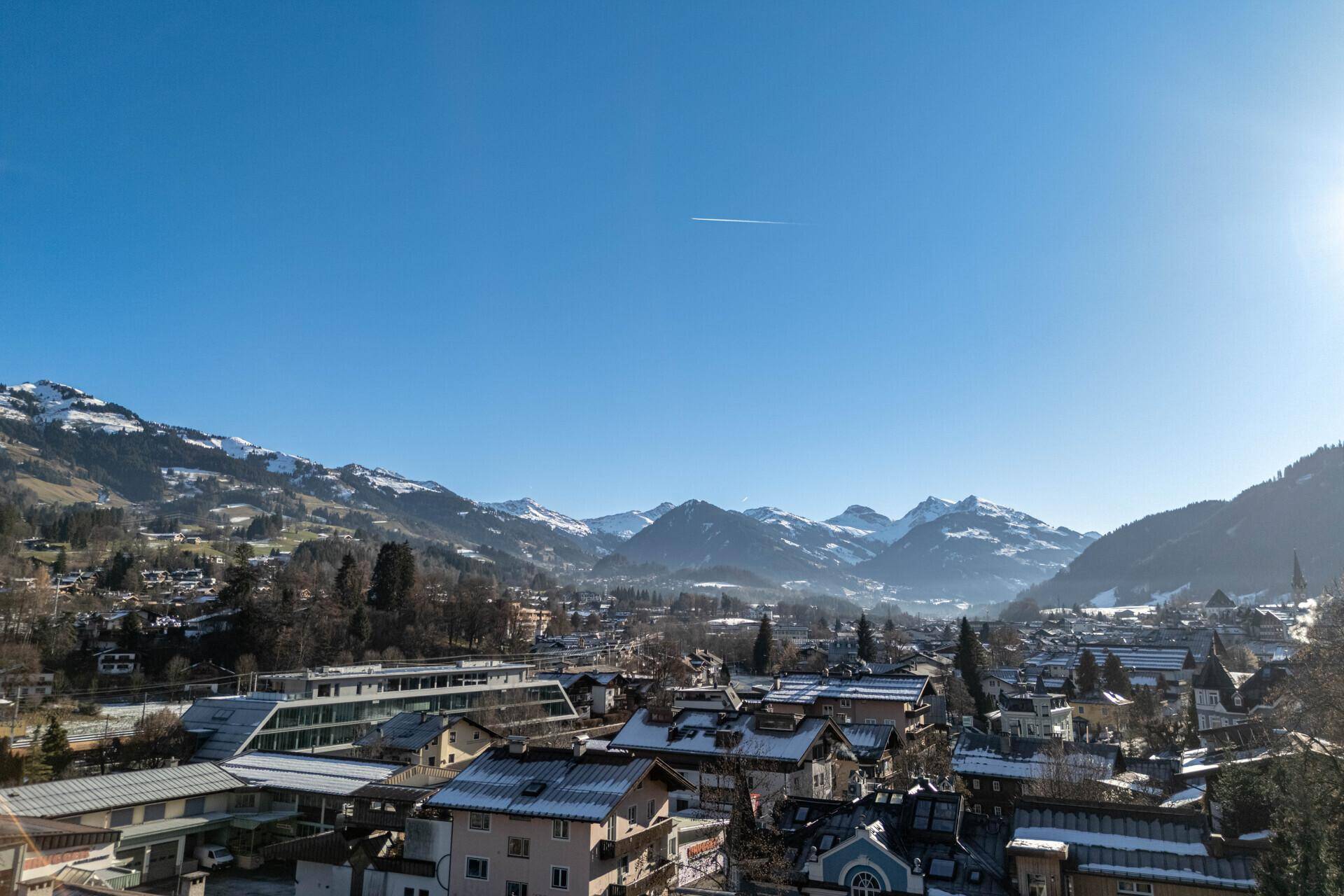 Panoramablick über eine verschneite Stadt im Tal, umgeben von majestätischen Bergen unter blauem Himmel.