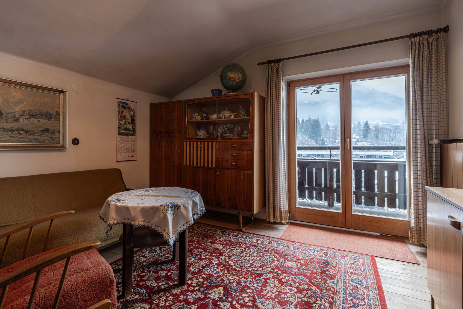 Gemütlicher Wohnraum mit traditionellen Möbeln, Teppichboden und Balkonzugang mit Bergblick.