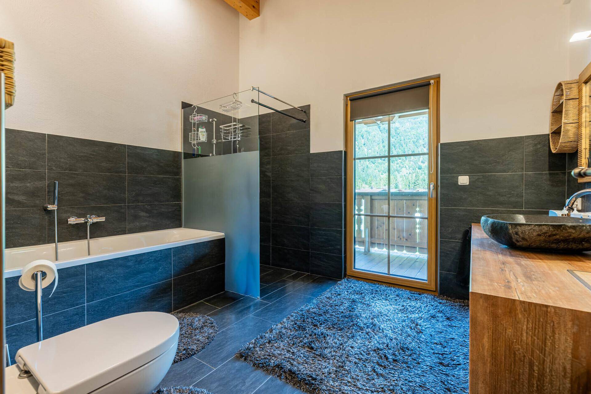 Modernes Badezimmer mit Badewanne, Dusche, dunklen Fliesen und Balkonzugang.