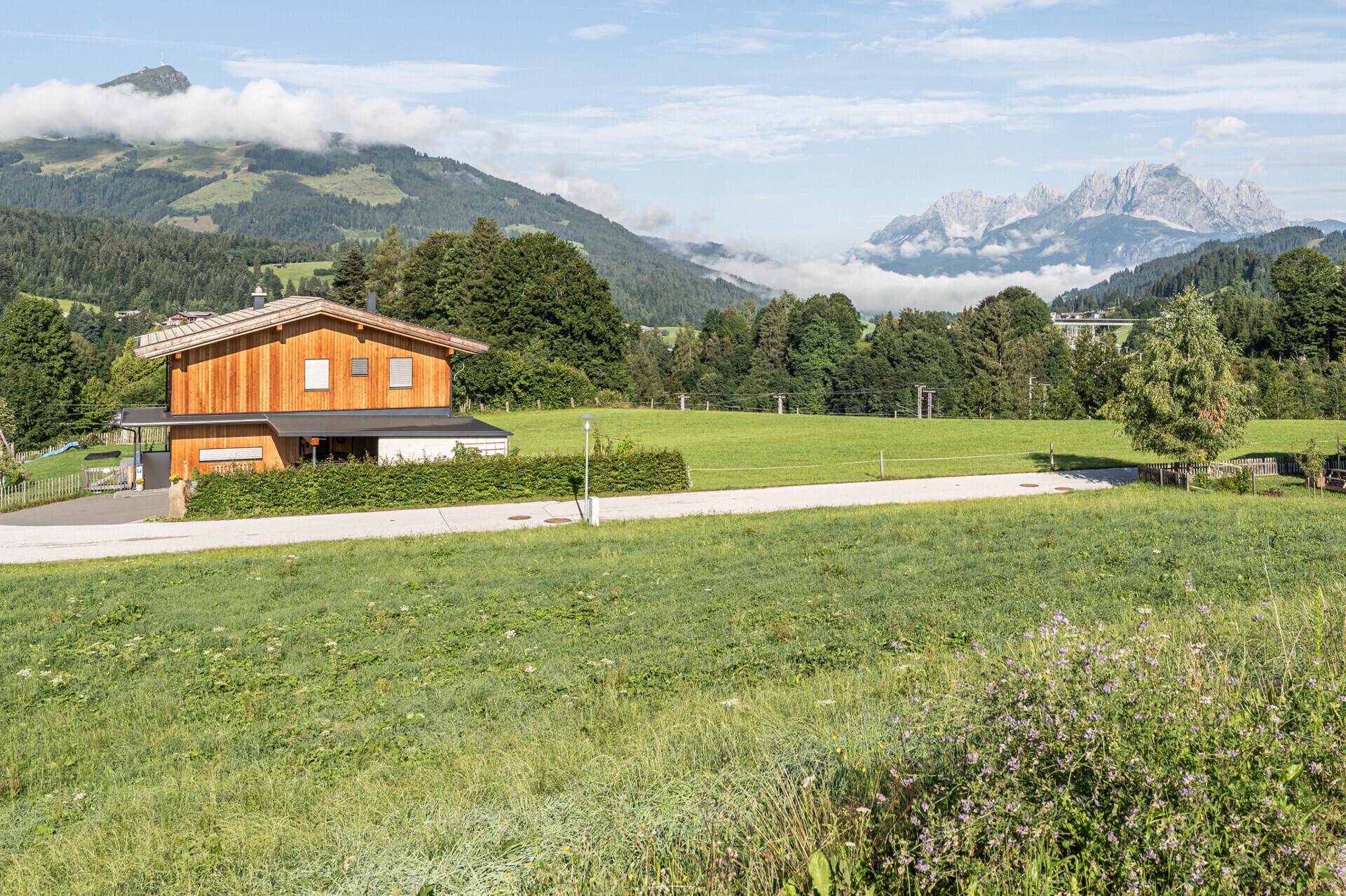 Modernes Holzhaus mit gepflegter Hecke und weitem Blick auf die umliegende Bergwelt unter blauem Himmel.