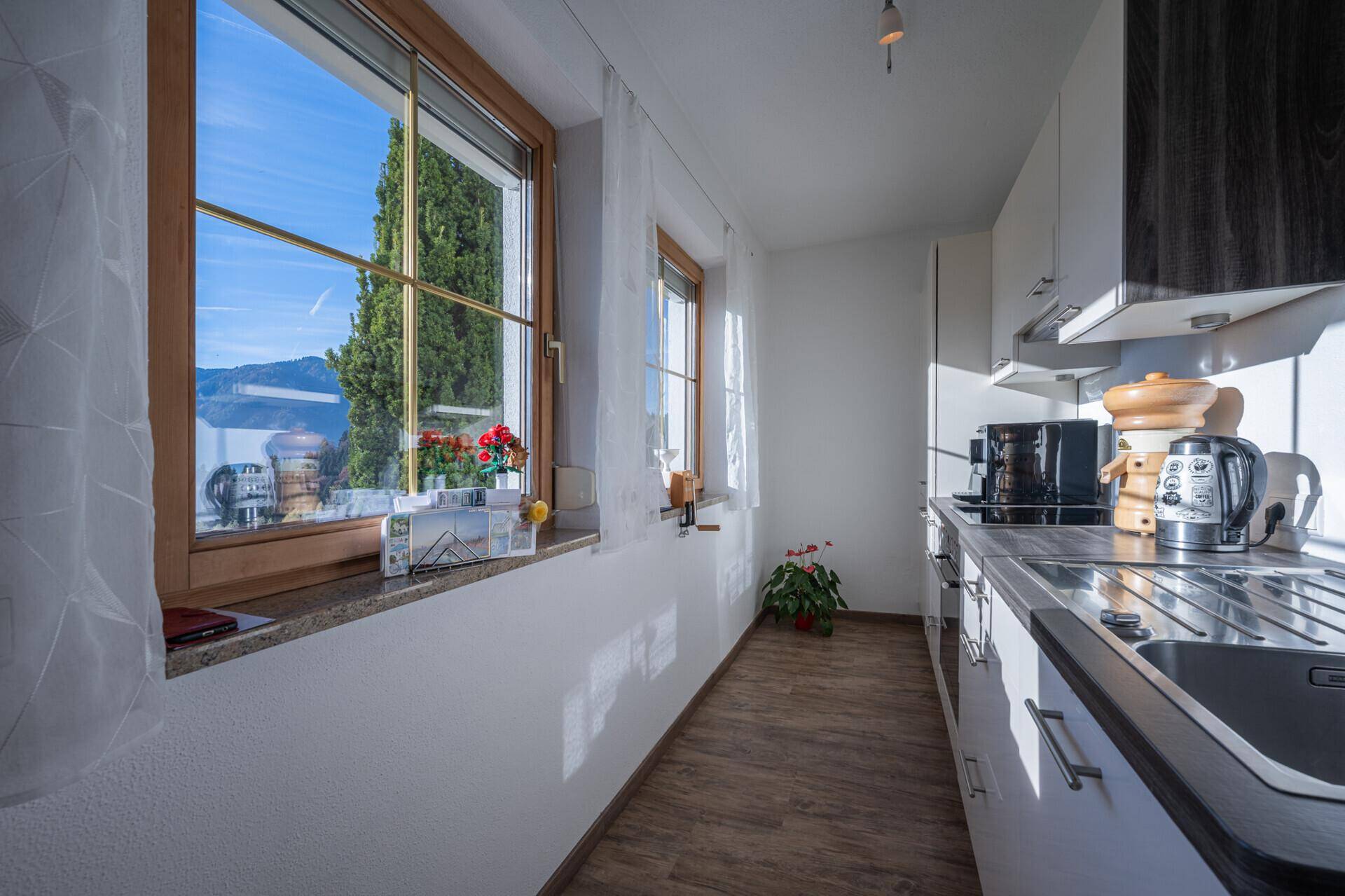 Schmale Küche mit weißen Schränken, Holzboden und Fenster mit Blick ins Grüne.