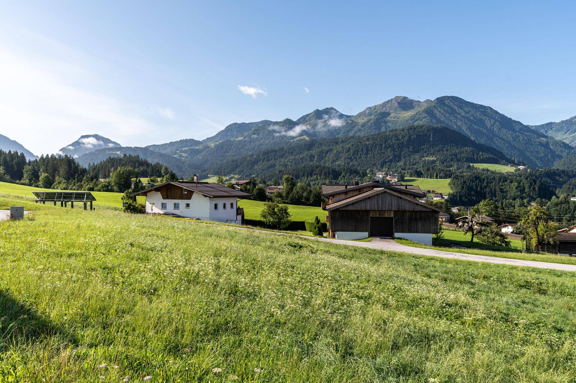 Ländliche Gebäude in einer weiten grünen Landschaft mit bewaldeten Bergen und Solaranlagen im Hintergrund.