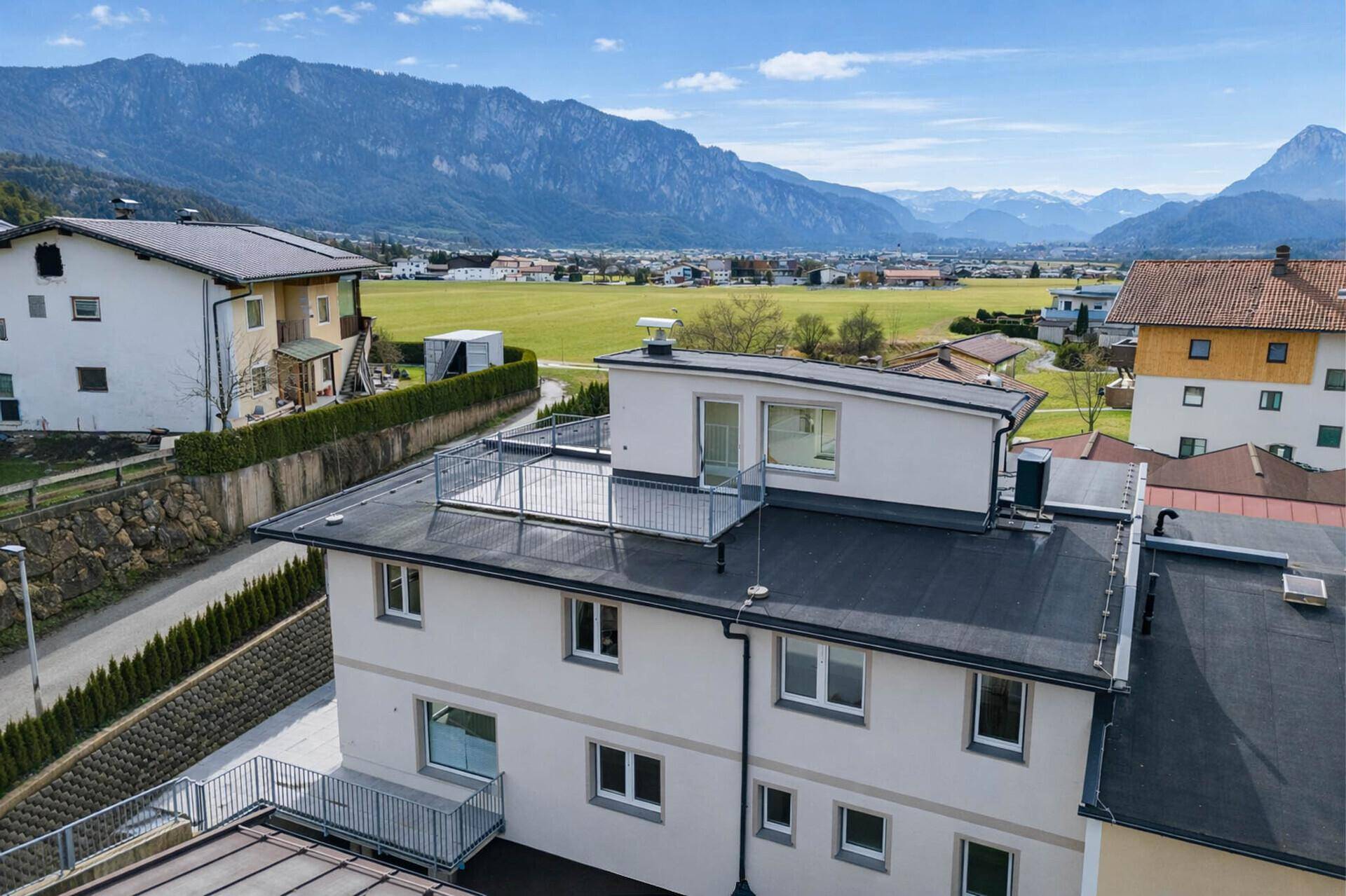 Gebäudeansicht mit Dachterrasse, umgeben von Feldern und einer beeindruckenden Bergkulisse im Hintergrund.