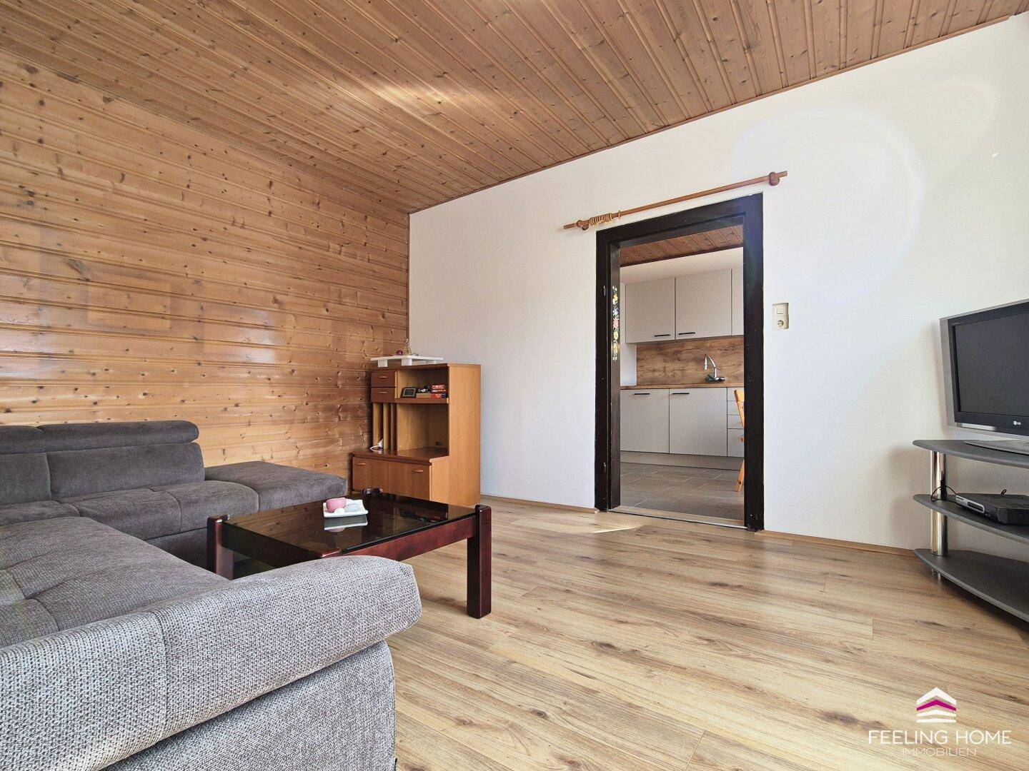 Wohnzimmer mit rustikaler Holzwandverkleidung, Parkettboden und offenem Zugang zur Küche.