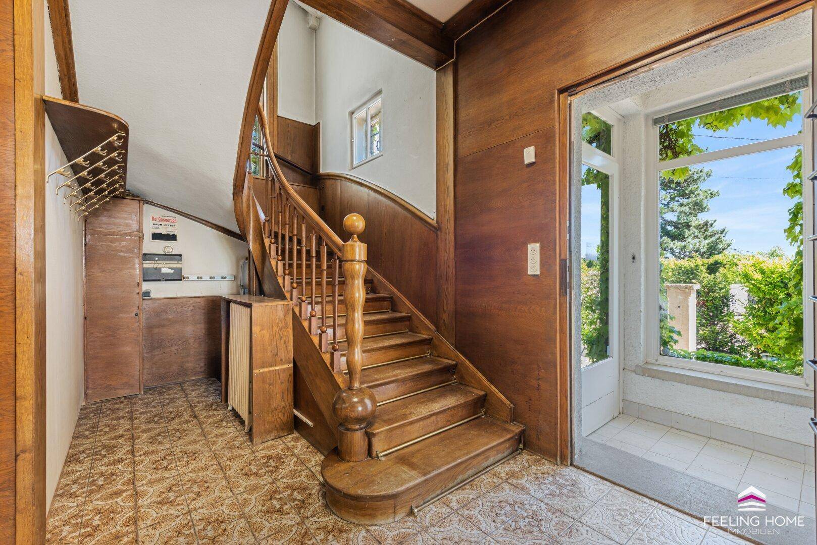 Eingangsbereich mit eleganter Holztreppe, Fliesenboden und viel Tageslicht durch die große Tür.