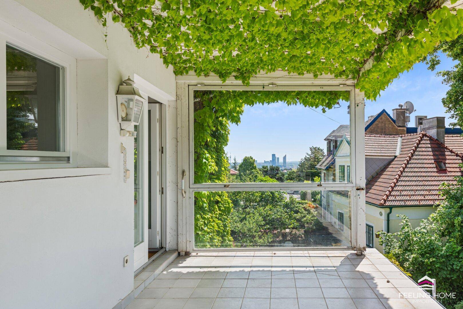 Überdachter Balkon mit Fliesenboden, grüner Bepflanzung und Fernblick auf die Stadt.