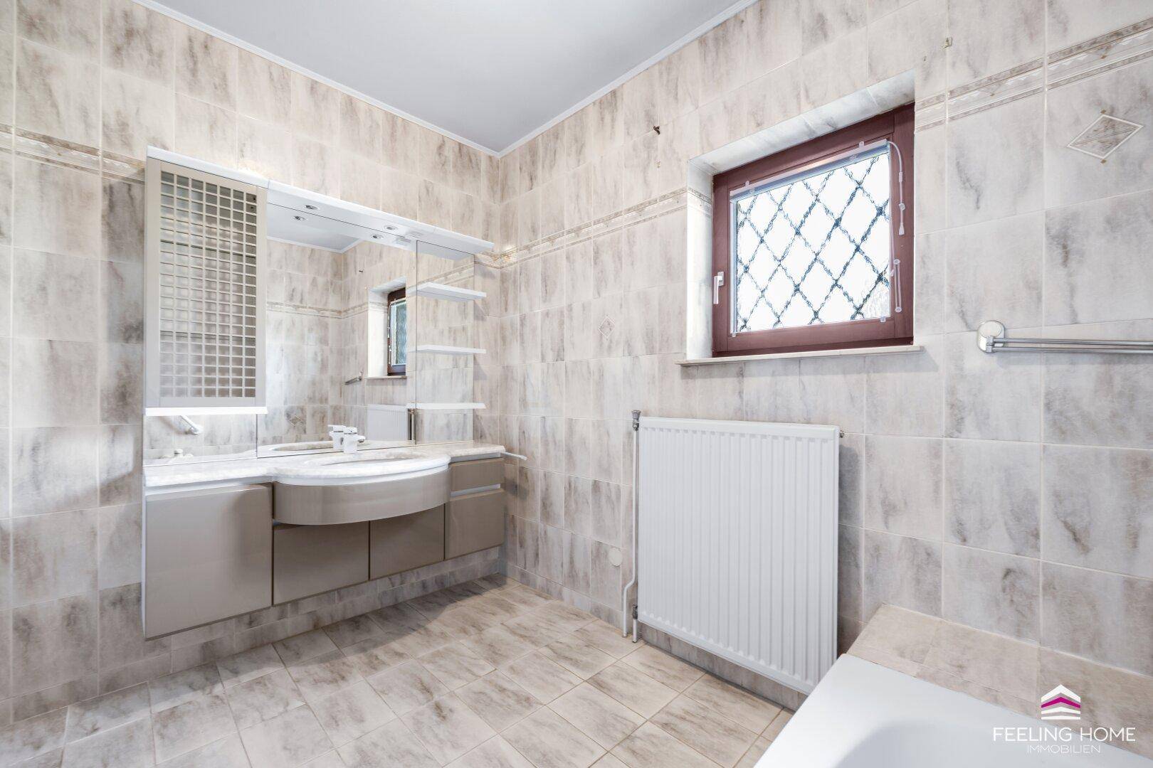 Gefliestes Badezimmer mit Doppelwaschbecken, Spiegelschrank, Badewanne und Fenster.