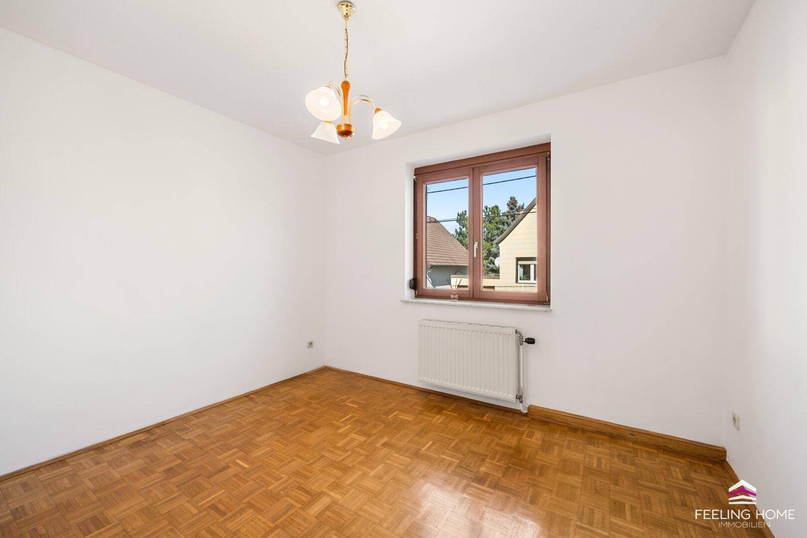 Schlafzimmer mit Parkettboden, Heizkörper und einem Fenster mit Blick ins Grüne.
