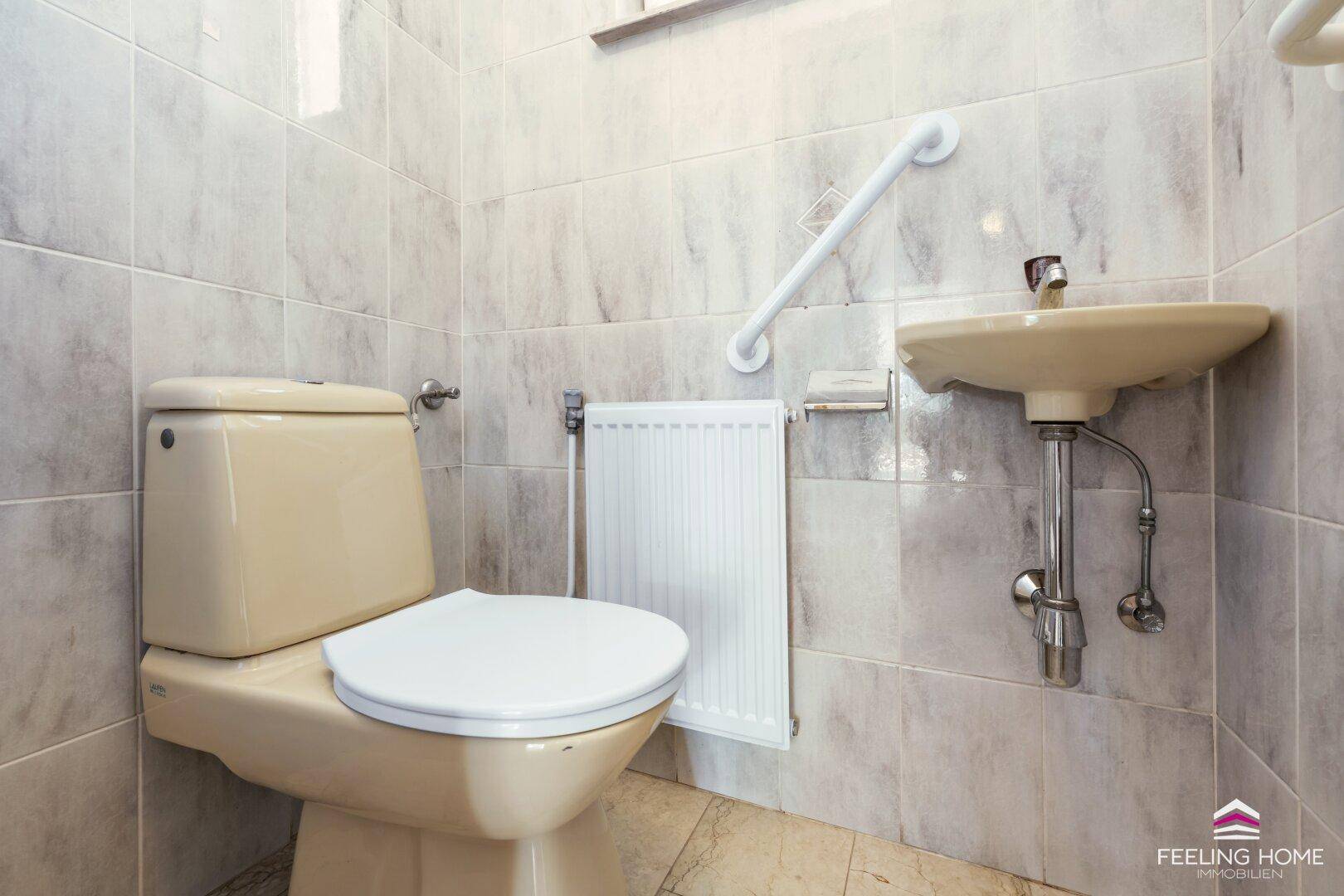 Kleines WC mit beigefarbenen Fliesen, Waschbecken und Haltegriff für Barrierefreiheit.